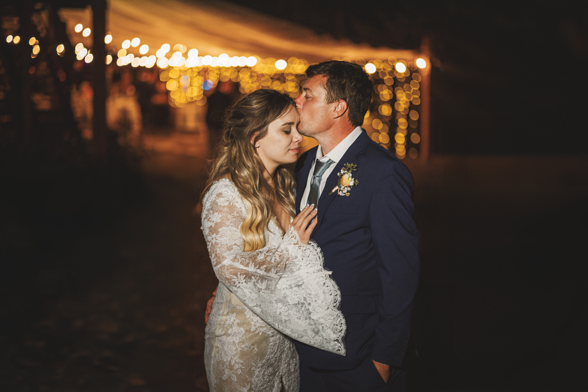 A bride and groom embrace under warm string lights, resembling a scene from Auto Draft. The groom tenderly kisses the bride's forehead. She stuns in her lace dress, while he looks sharp in his suit with a boutonniere.