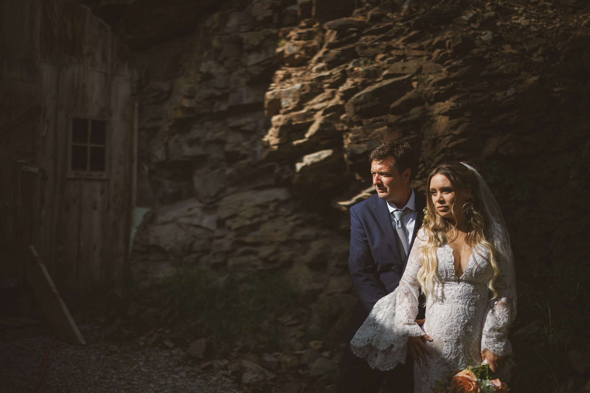 A groom in a navy suit and a bride in a white lace dress with long sleeves stand close, holding hands, against a rocky background with sunlight partially illuminating them—like an auto draft of nature's perfect moment.