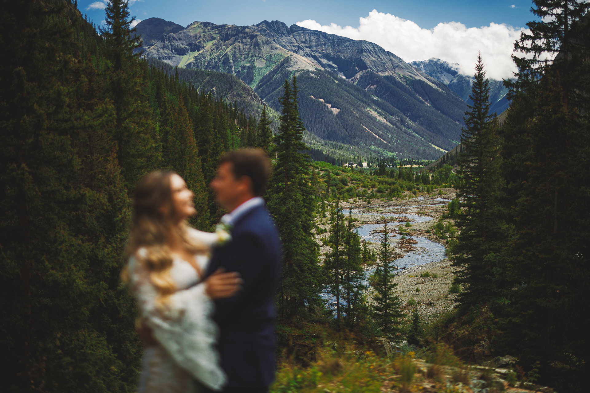 A blurred couple stands embracing with a scenic mountain landscape, forest, and river in the background under a partly cloudy sky, creating an image that could easily be an auto draft of nature's splendor.