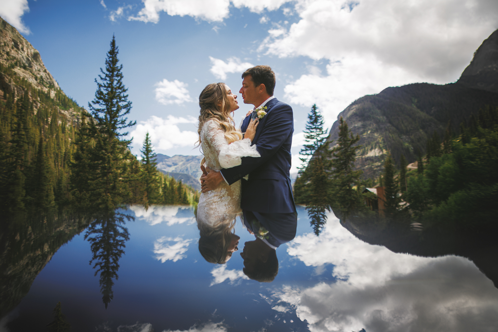 A bride and groom embrace by a reflective water surface amidst a mountainous landscape with pine trees, under a partly cloudy sky. The scene feels almost like an Auto Draft of nature's most breathtaking elements.