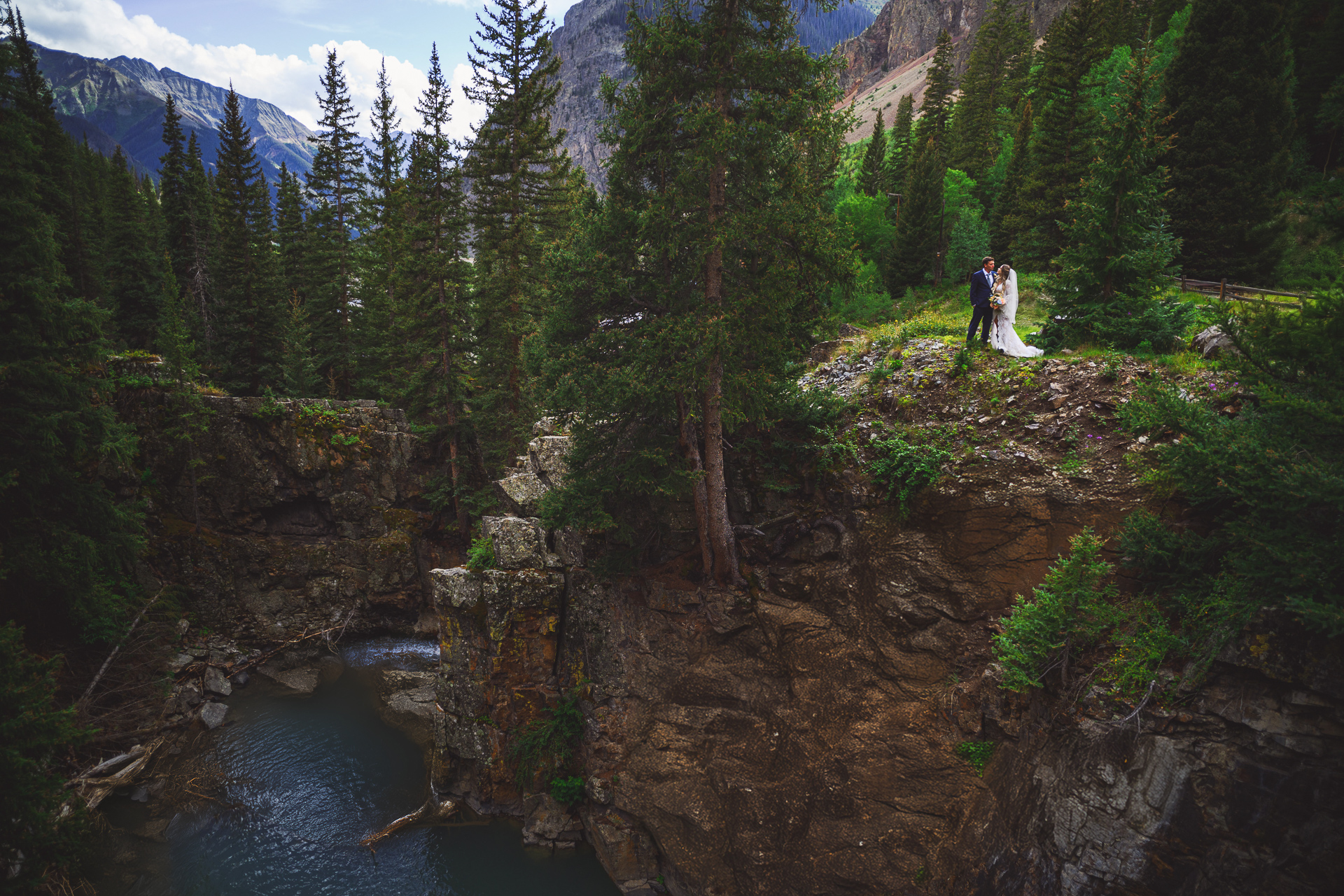 Bride and groom stand on a rocky cliff surrounded by trees, Auto Draft capturing their moment as they overlook a small waterfall and pool of water in a mountainous landscape.