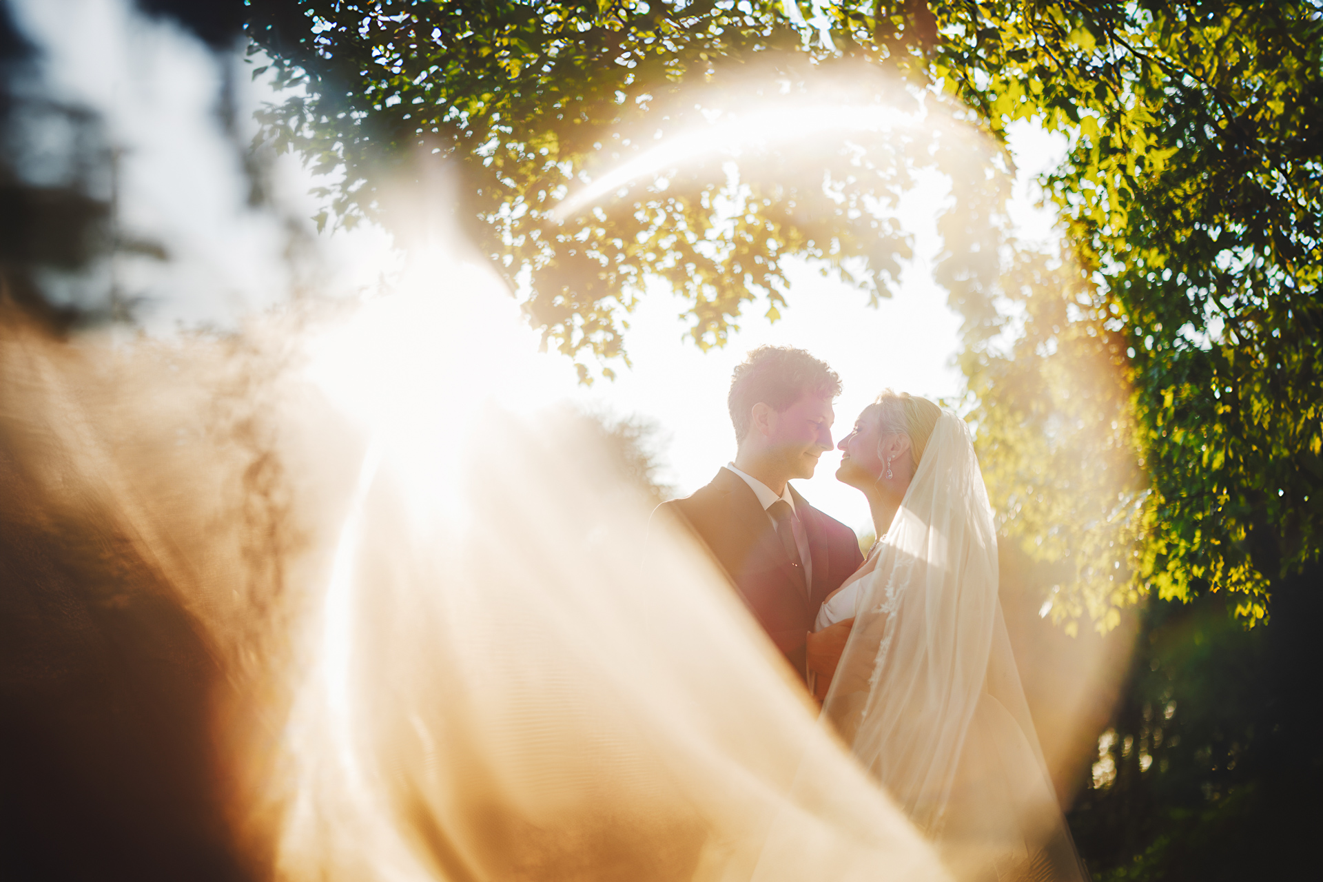 A couple stands together outdoors in wedding attire, with sunlight creating a glowing, circular effect around them, like an auto draft of a perfect moment. Trees and foliage are visible in the background.
