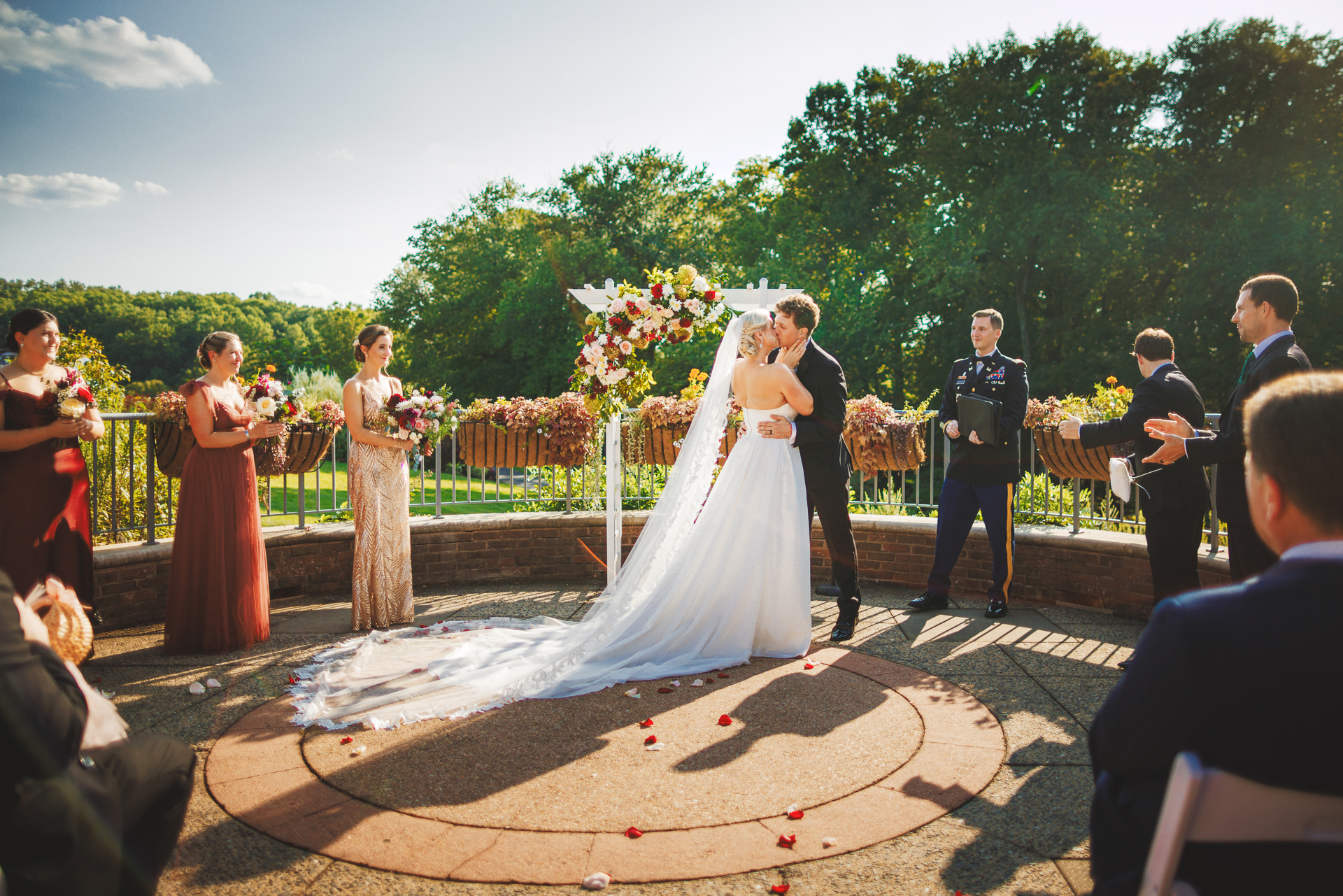 A couple kisses at an outdoor wedding ceremony, Auto Draft capturing the moment perfectly. They are surrounded by their wedding party and guests, with a floral arch in the background and trees in the distance.