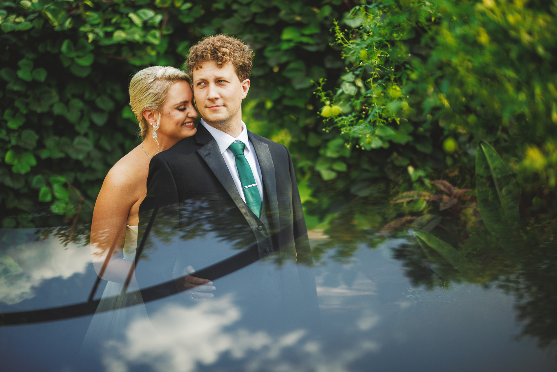 A couple, dressed in formal attire, stands closely together in front of lush greenery. The woman leans her head on the man's shoulder, both looking content as if their love story just came right out of an auto draft fairy tale.