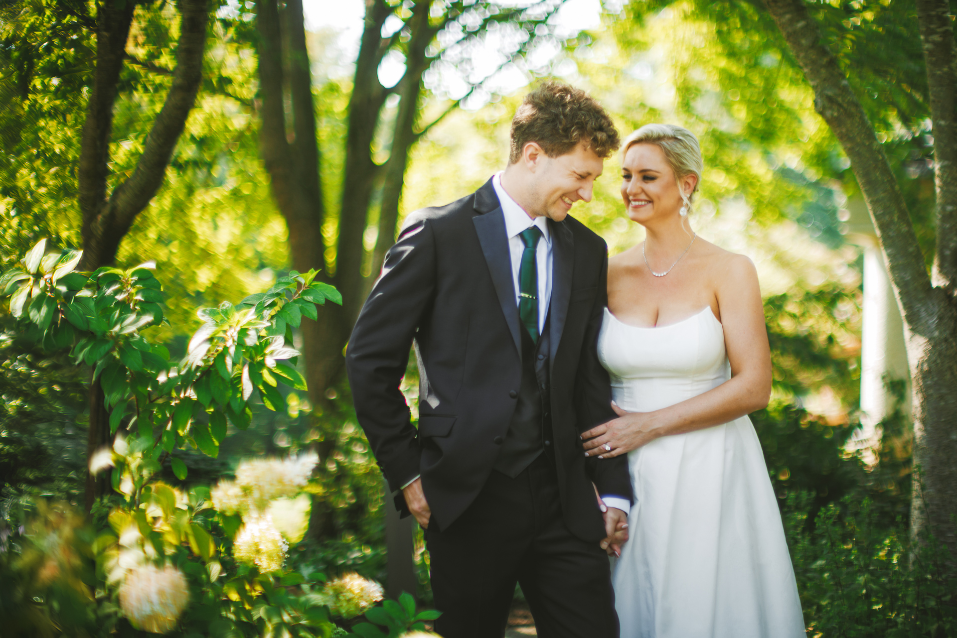 A couple in formal attire, with the man in a suit and the woman in a white dress, walk hand in hand outdoors amidst lush greenery. They both appear to be smiling, as if creating their own auto draft of cherished memories.