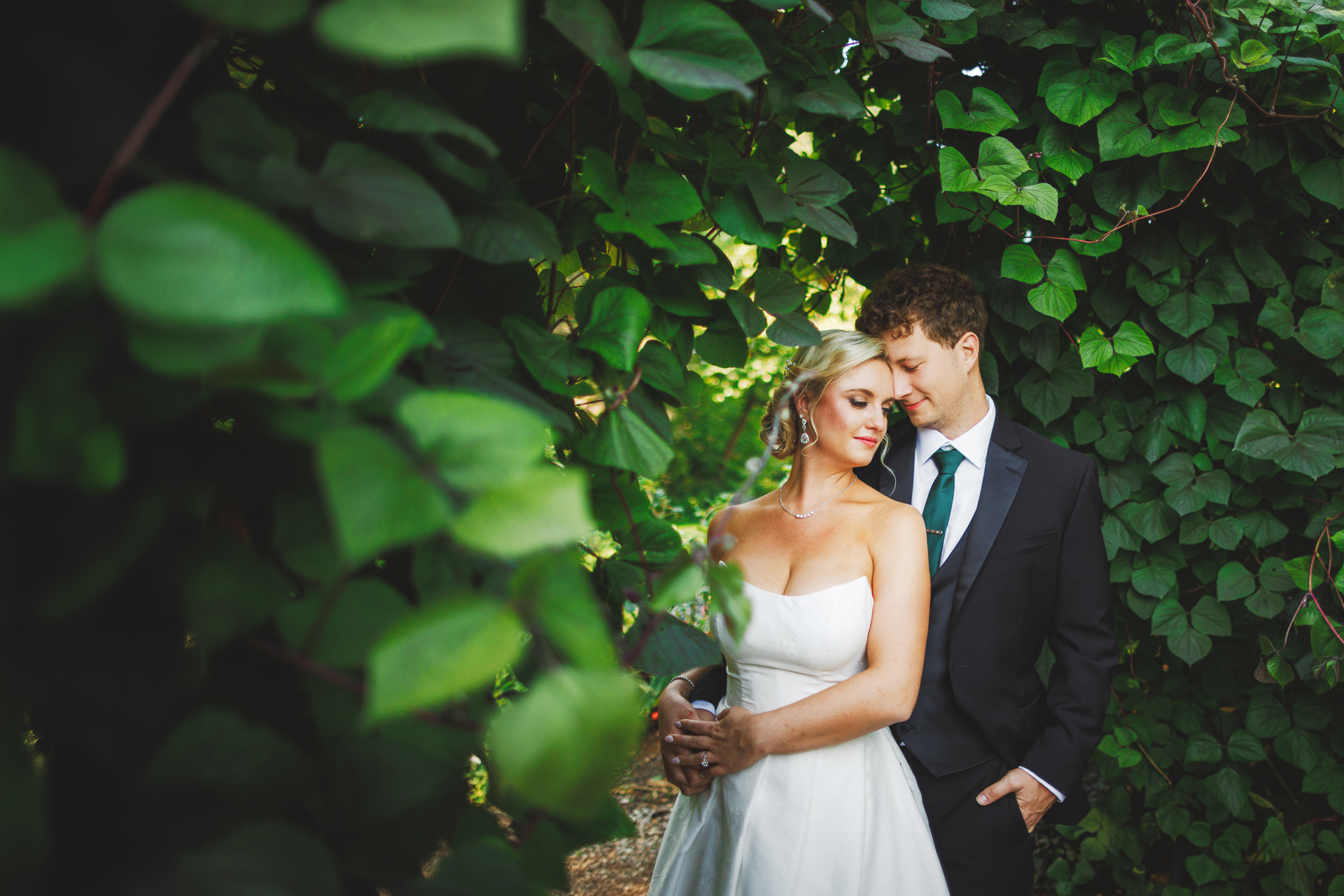 A bride and groom stand closely together in a lush green garden, with the groom gently resting his head on the bride's, capturing a moment so perfect it seems like an Auto Draft of love.