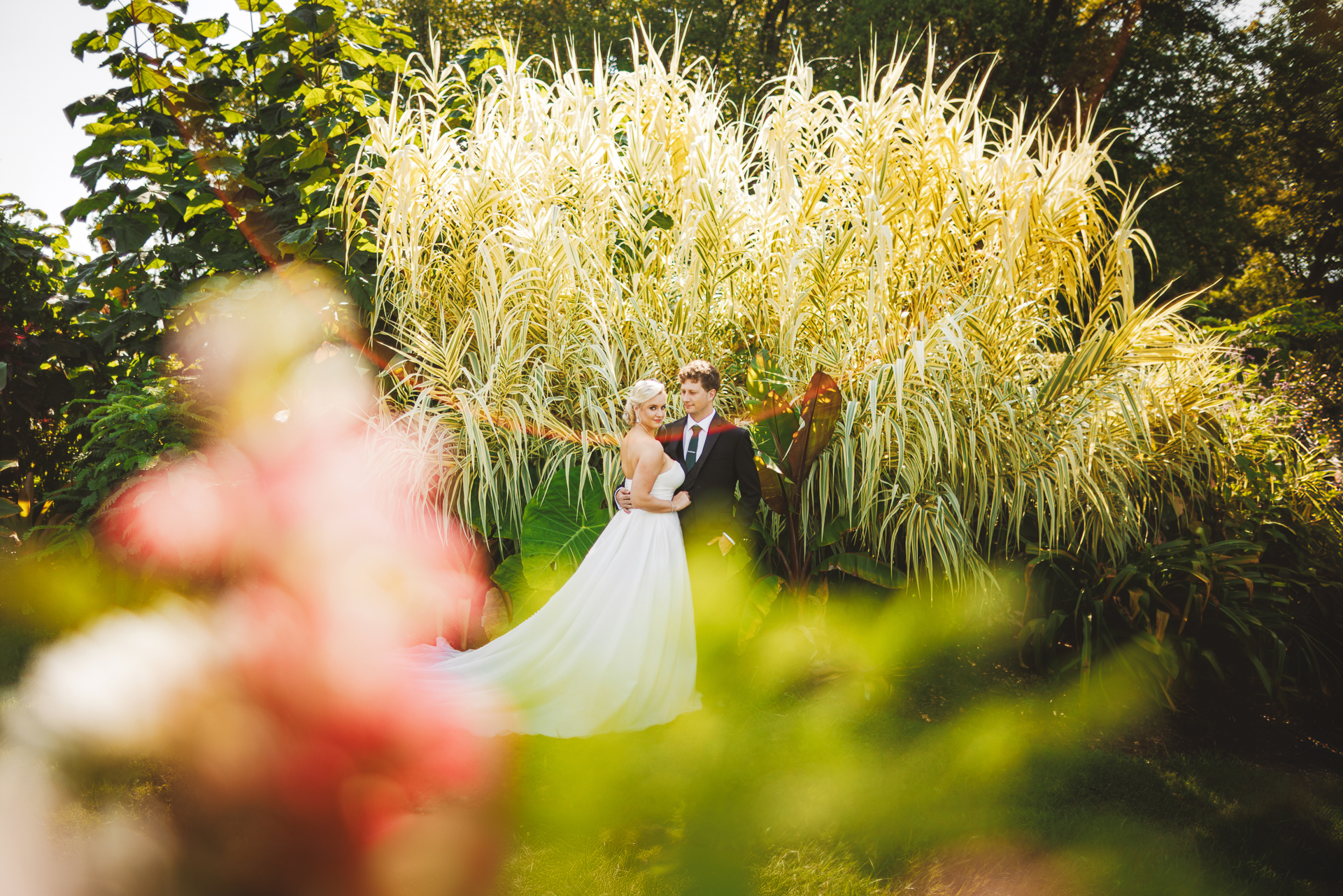 A bride and groom stand in front of tall, green foliage, posing for a wedding photo in an outdoor garden setting, with moments seemingly captured in an Auto Draft of perfection.