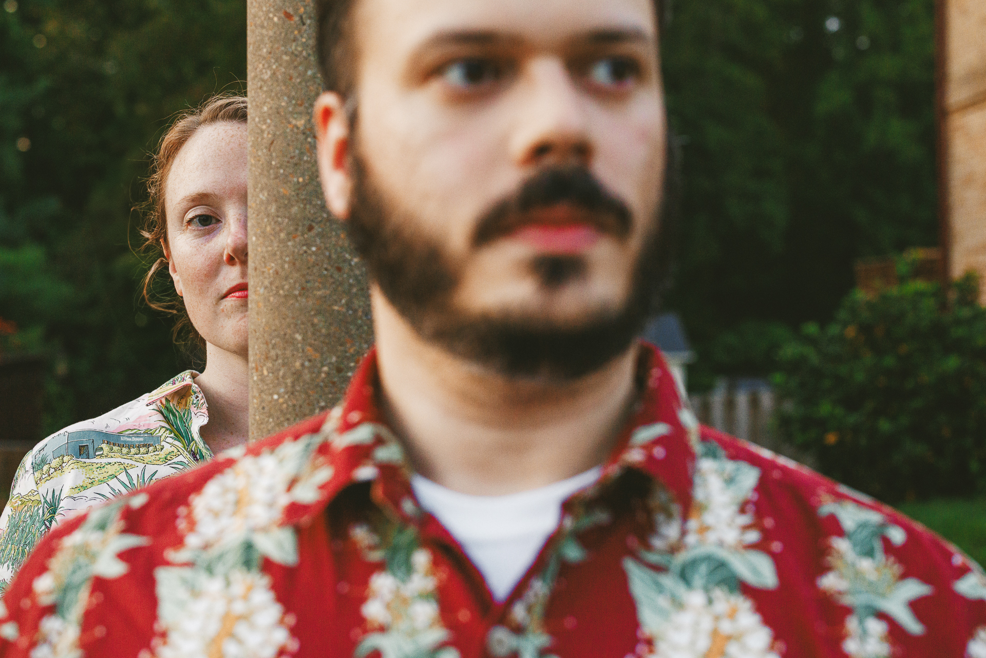 Two people in floral shirts stand outside, partially obscured by a vertical pole; one is in the foreground, the other's face mostly hidden. Green foliage and a building are visible in the background, like an artistic auto draft capturing an unscripted moment.