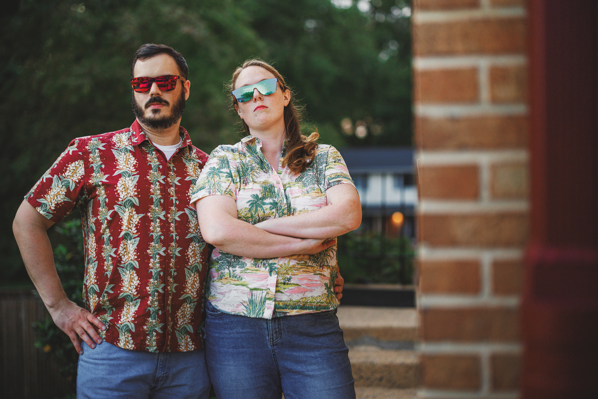 Two adults wearing colorful Hawaiian shirts and mirrored sunglasses stand outdoors, one with arms crossed, both looking directly at the camera with serious expressions. Against the backdrop of a brick wall and greenery, they seem like an auto draft from a stylish vacation magazine.