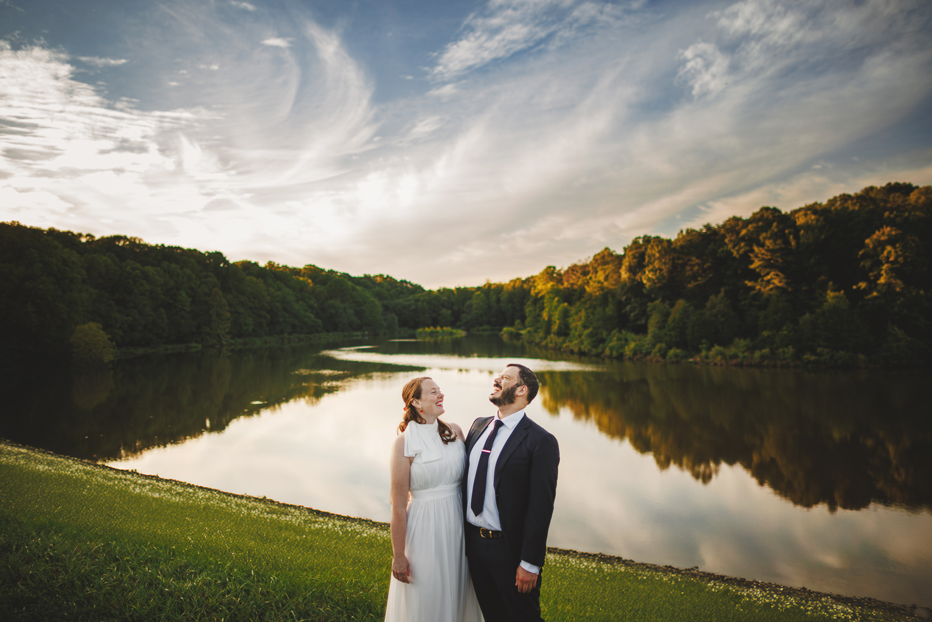 A couple stands by a calm lake with a forested shoreline under a partly cloudy sky. Both individuals are smiling and looking upward, as if an auto draft of nature’s beauty is being reflected perfectly in the tranquil water below.