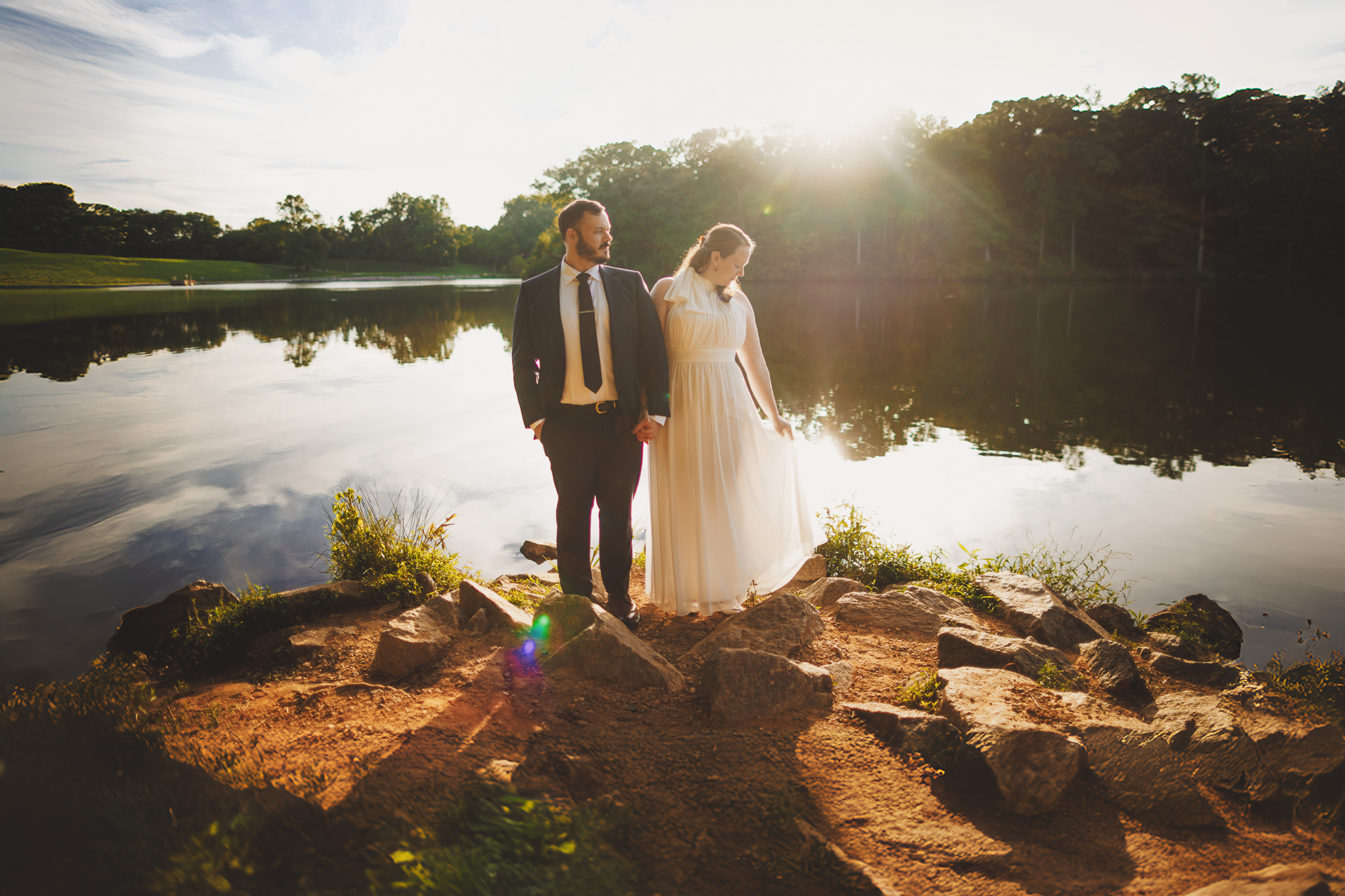 A couple, dressed in a suit and a white dress, stand hand in hand on a rocky shoreline by a lake as the sun sets in the background, capturing their perfect moment like an auto draft of love's first light.