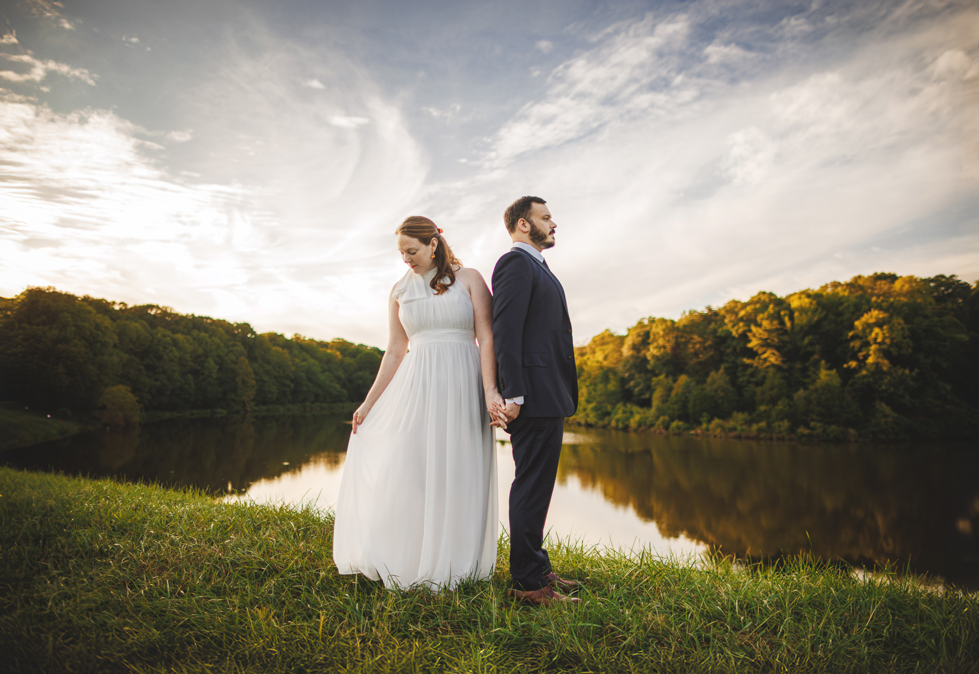 A bride and groom stand back-to-back holding hands in front of a serene lakeside and forested background under a partly cloudy sky, as if the scene was flawlessly captured by an auto draft.