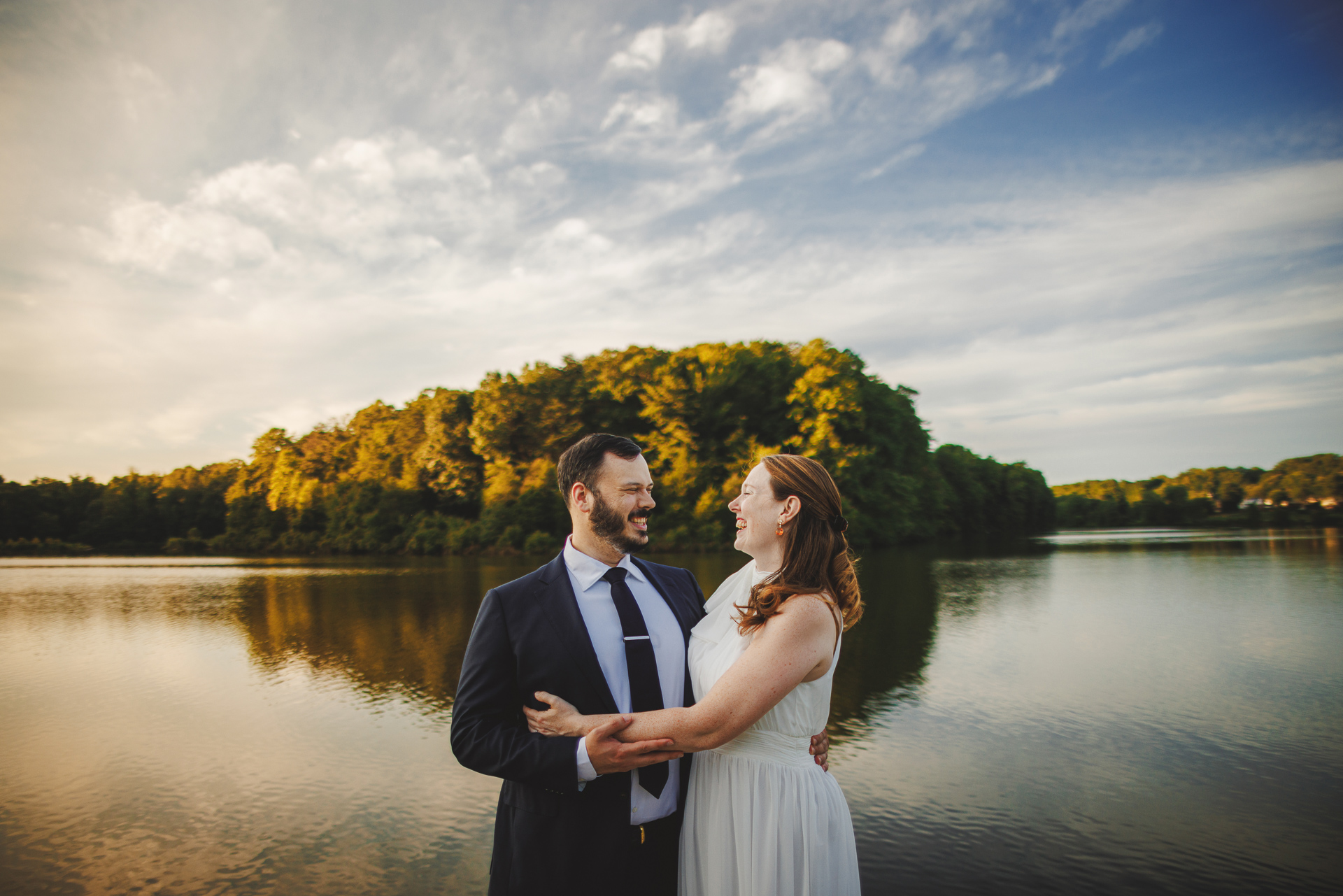 A couple stands in front of a serene lake, embraced and smiling at each other, with a lush, tree-covered shore and a blue sky with clouds in the background. The scene feels like an auto draft of nature's perfect moment.