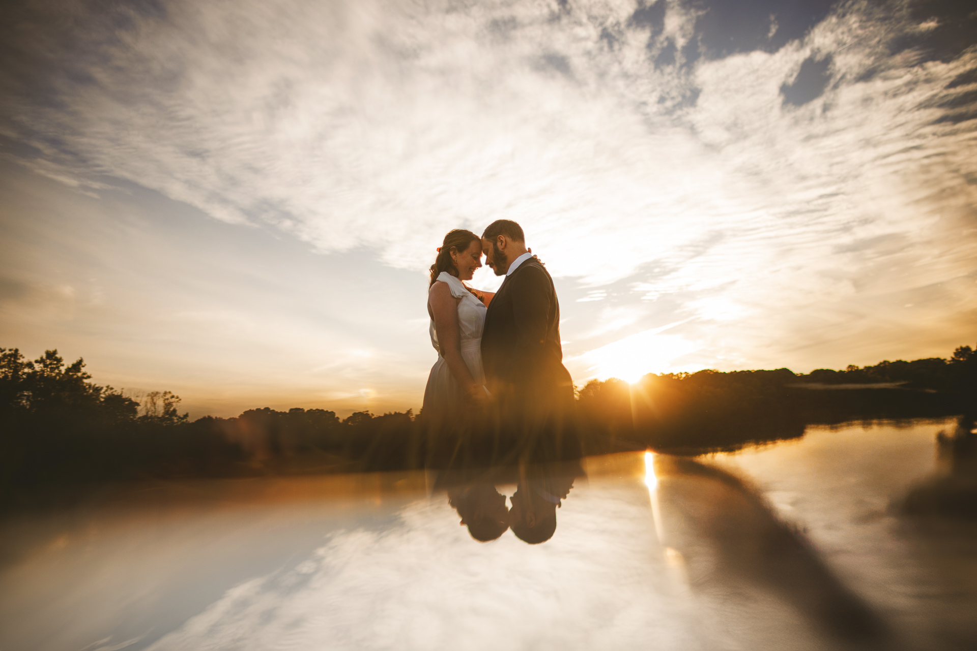 Silhouetted couple in formal attire face each other at sunset by a reflective body of water, with a cloudy sky creating a warm, serene backdrop. The moment feels like an auto draft of nature's perfect evening sketch.