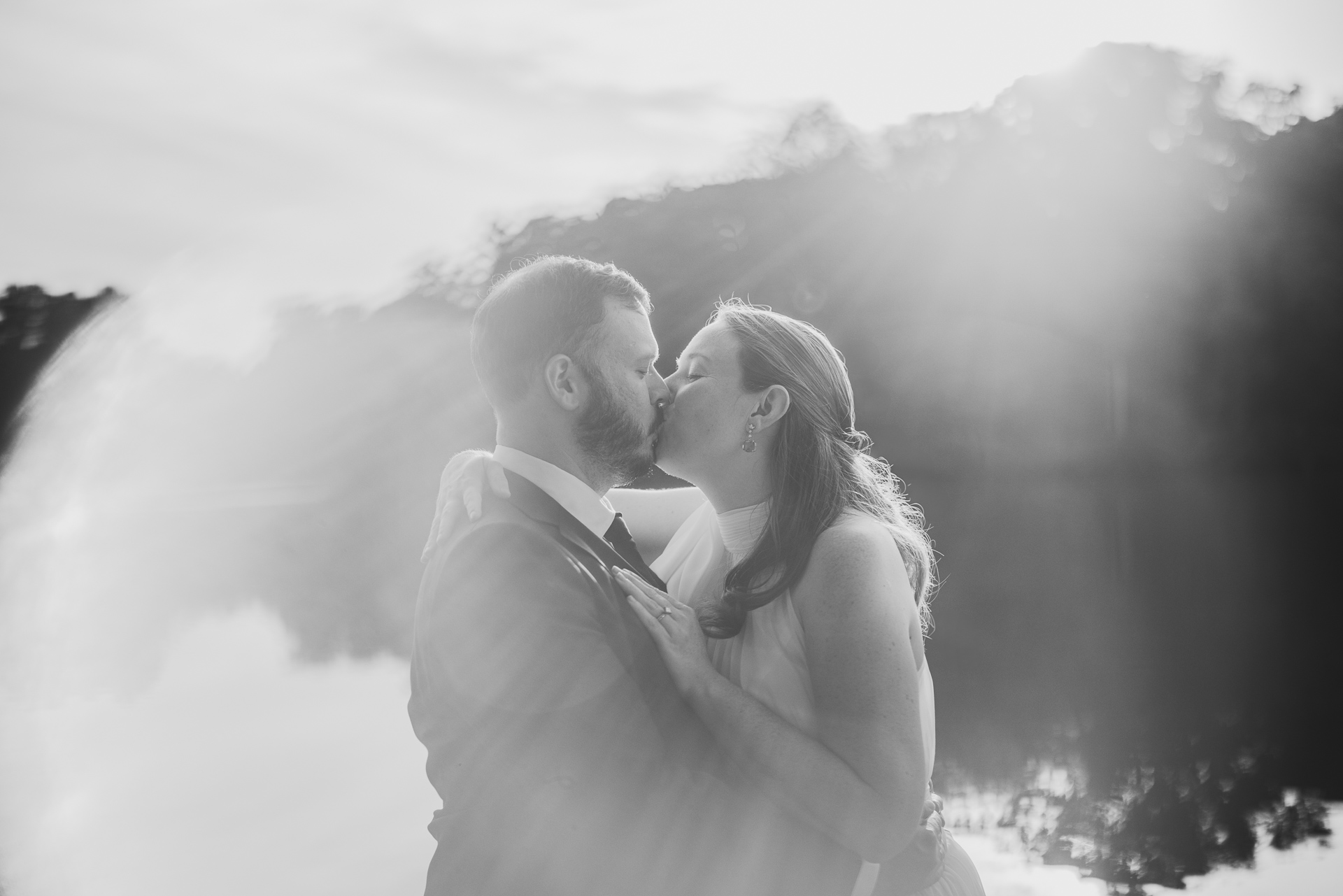 A couple kisses by a calm lake, surrounded by rays of sunlight, with trees in the background. Black and white Auto Draft photograph.