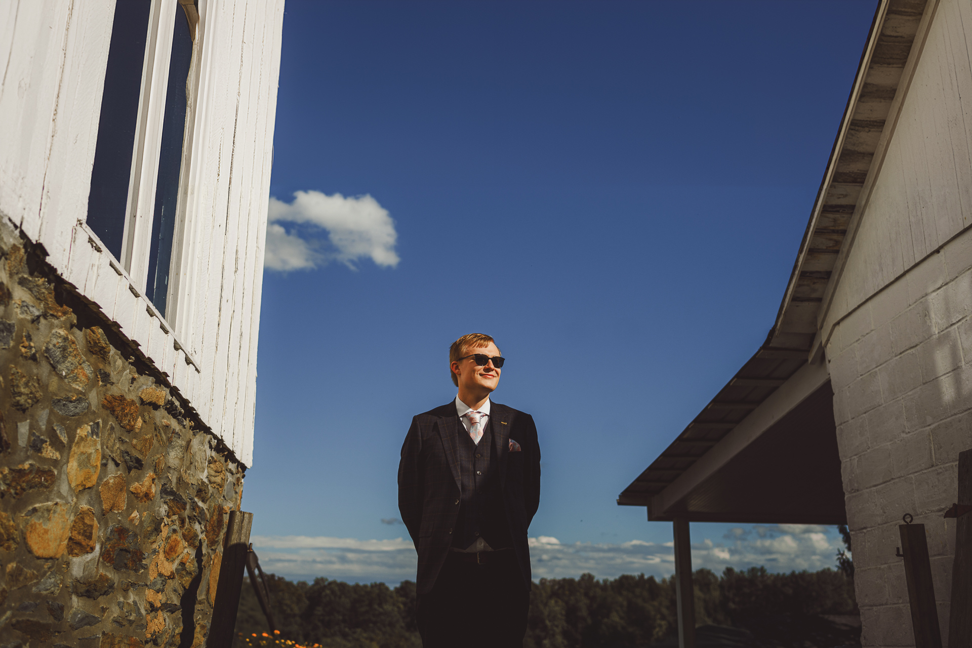 Man in a dark suit and sunglasses stands between two buildings against a bright blue sky, as if he just stepped out of an auto draft.