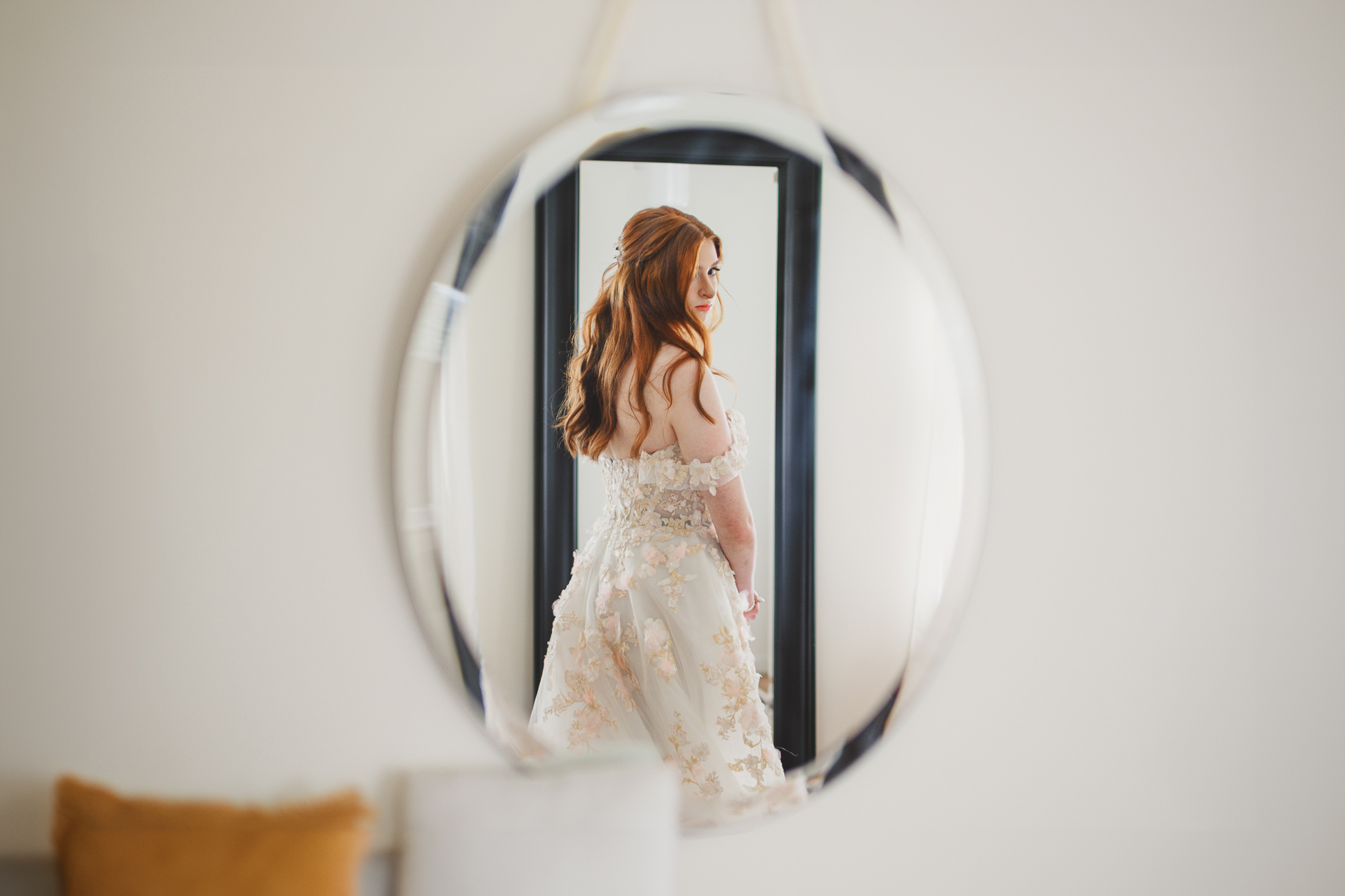 A bride in an off-shoulder wedding dress, reflected in a round mirror, meticulously prepares for her wedding day with the precision of an auto draft.