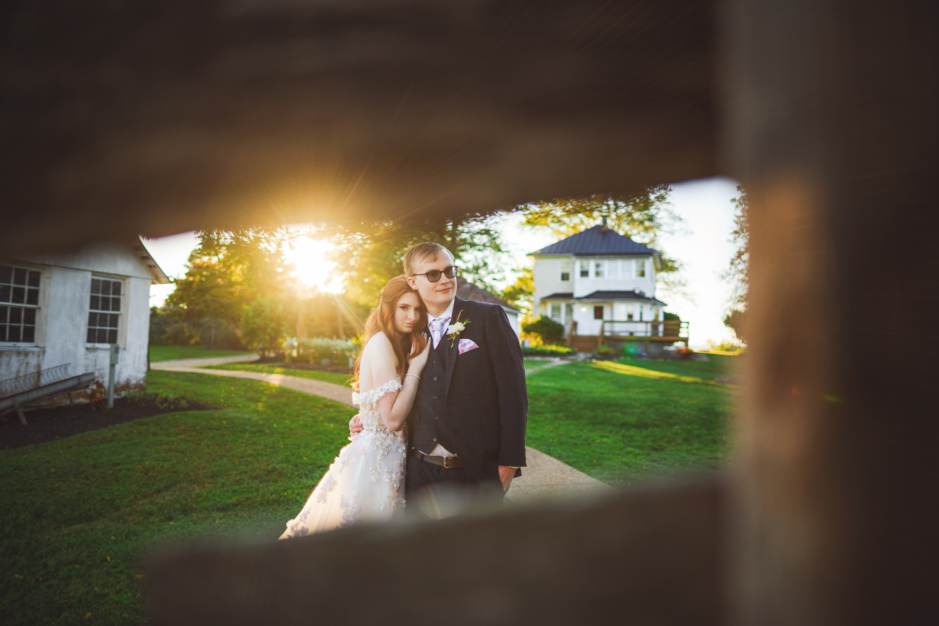 A couple in formal attire poses outdoors in front of a house, bathed in a warm glow from the sunlight behind them. The image is framed through a wooden fence, capturing an auto draft of their timeless moment.
