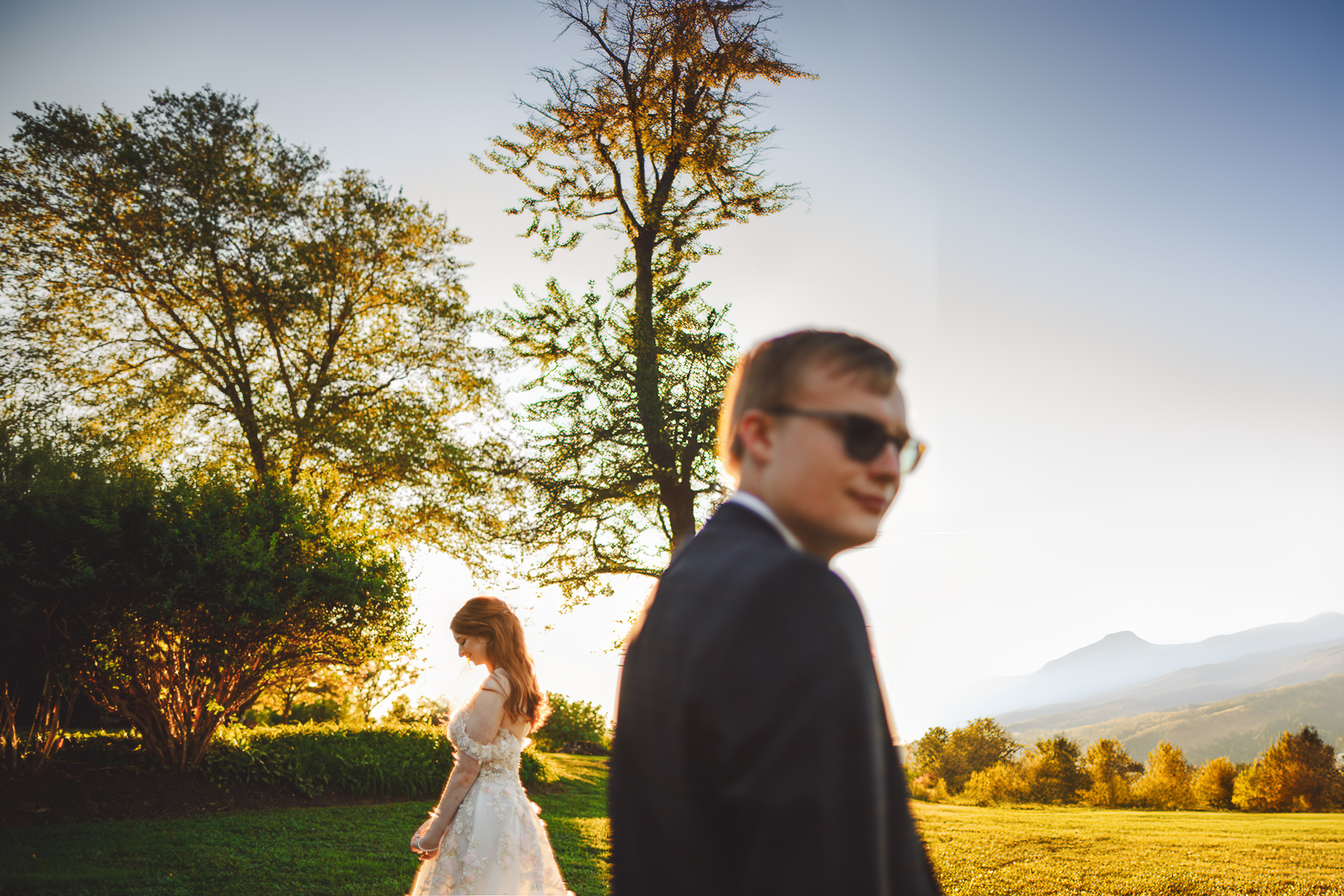 A person wearing sunglasses stands in the foreground while another person in a white dress, like an auto draft of beauty, stands in the background, surrounded by trees during sunset.
