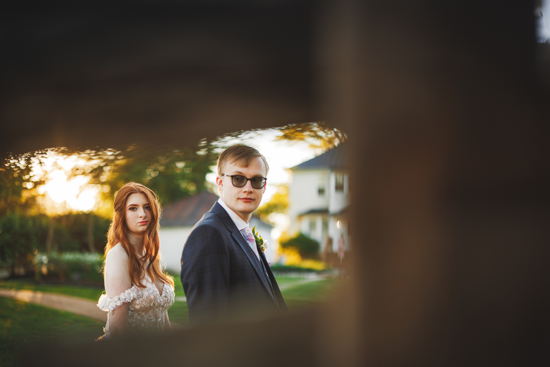A bride and groom stand outdoors, both looking towards the camera with a solemn expression. The background features greenery and a blurred view of a house, almost as if captured in an auto draft moment.