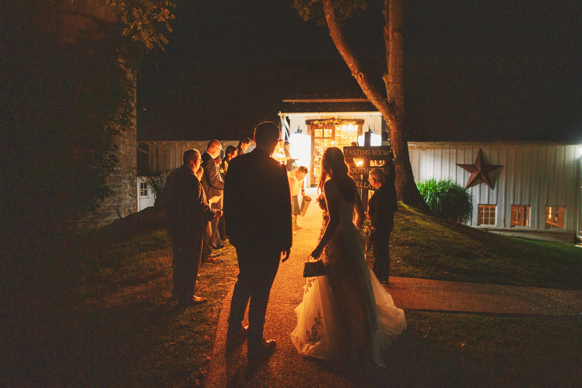 A bride and groom walk towards a lit building at night as guests look on, the perfect moment captured in an auto draft of memories.