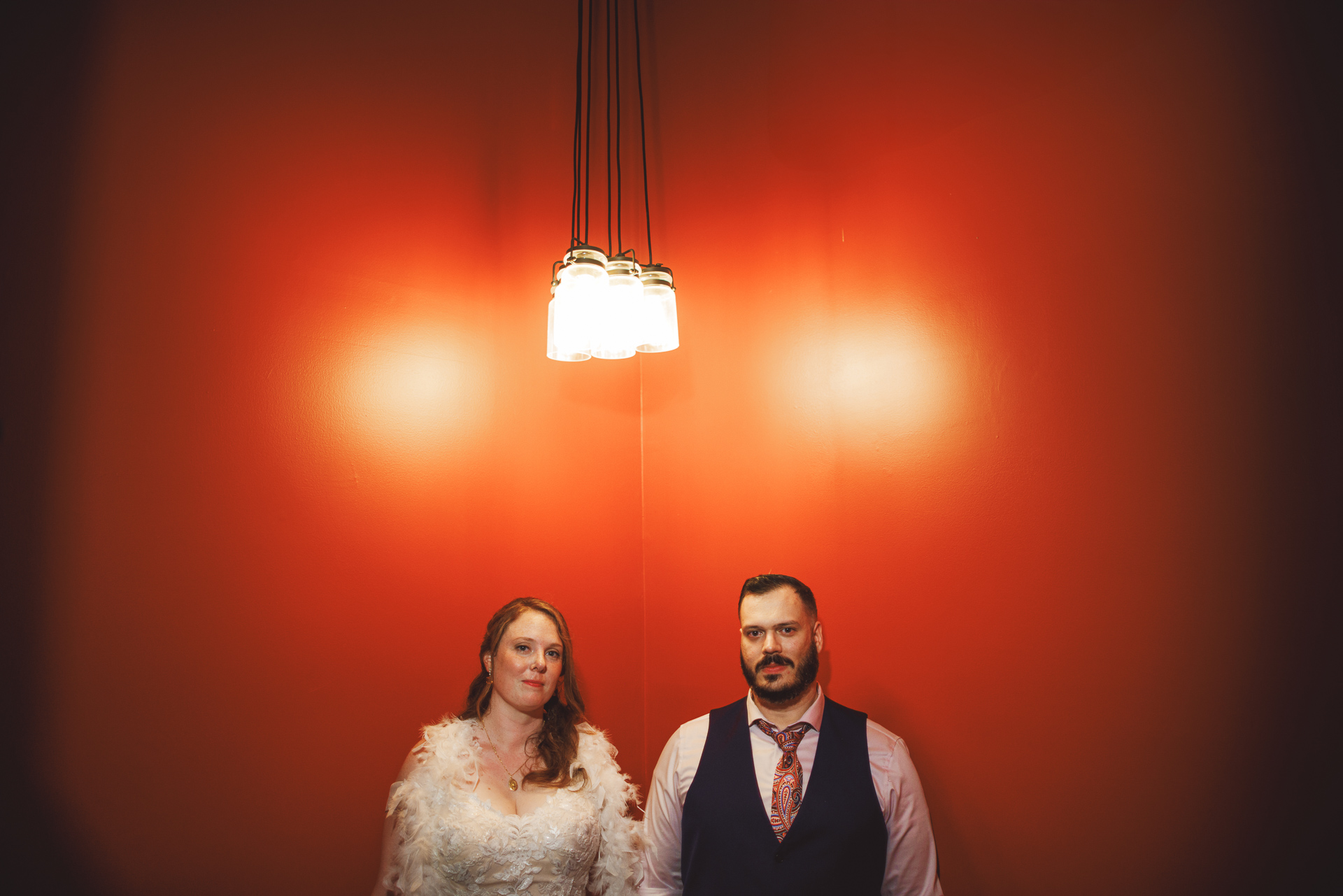A woman in a white dress and a man in a vest and tie stand side by side against a bright orange wall under hanging lights, their poised presence highlighted as if captured in an Auto Draft moment.