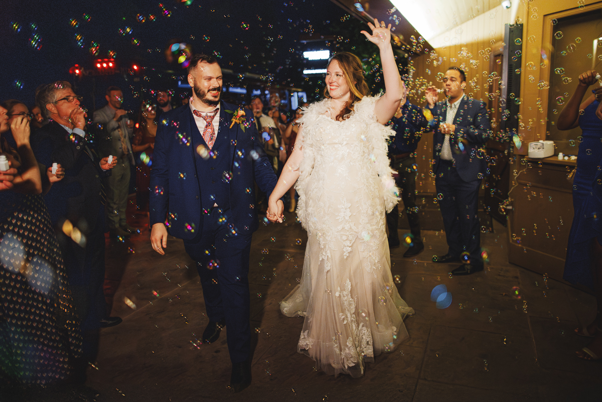 A bride and groom in formal wedding attire walk hand in hand through a crowd of guests. The bride waves while bubbles float around them, creating a whimsical scene that looks straight out of an auto draft fairy tale.