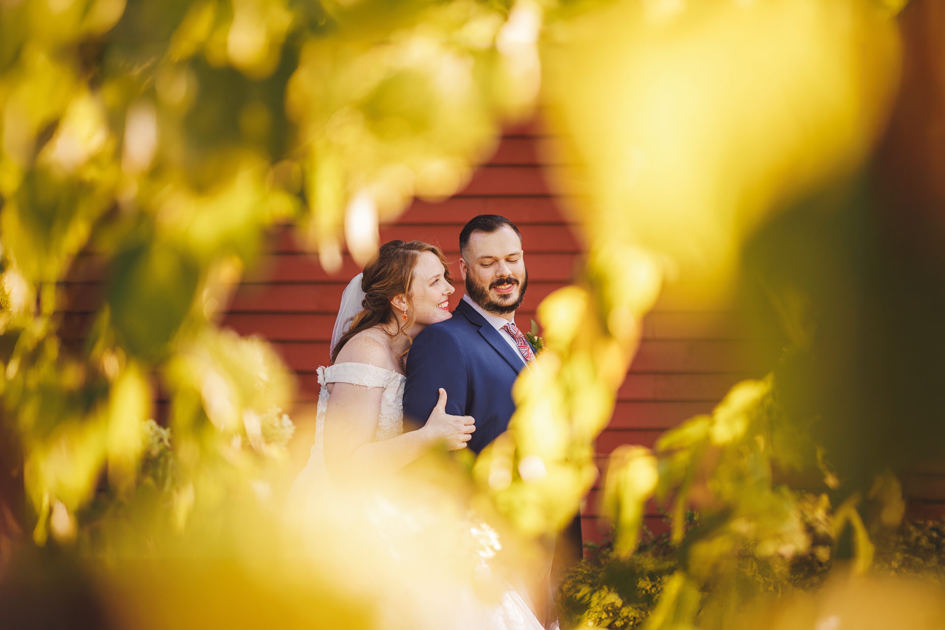 A bride and groom stand closely together, framed by yellow-green foliage, with a red wooden wall in the background, capturing an idyllic moment that seems as if it could be an auto draft of nature's perfect scene.
