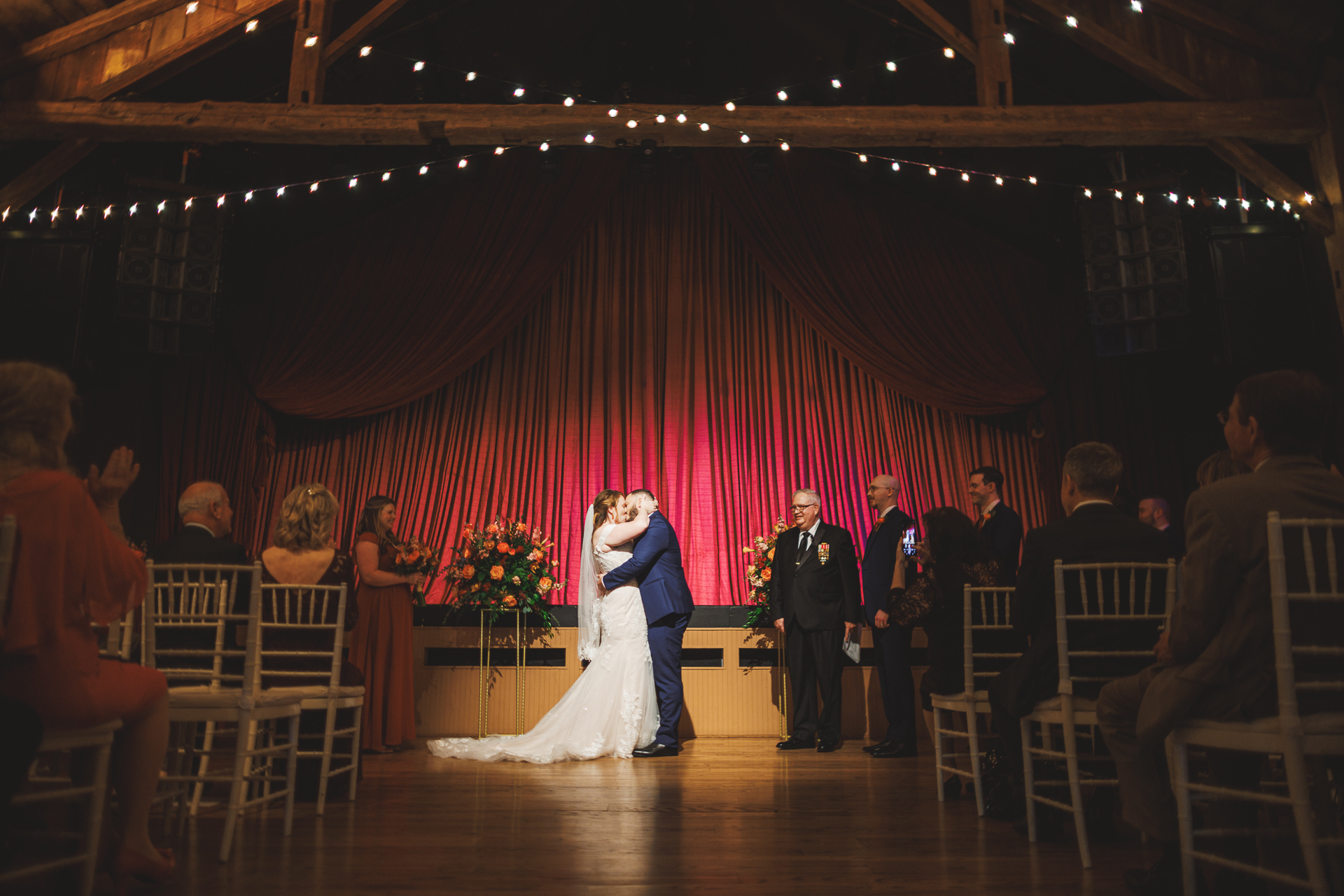 A couple in wedding attire shares a kiss on stage while guests seated in rows watch. String lights and draped red curtains decorate the background, creating a moment so perfect it could be straight out of an Auto Draft.