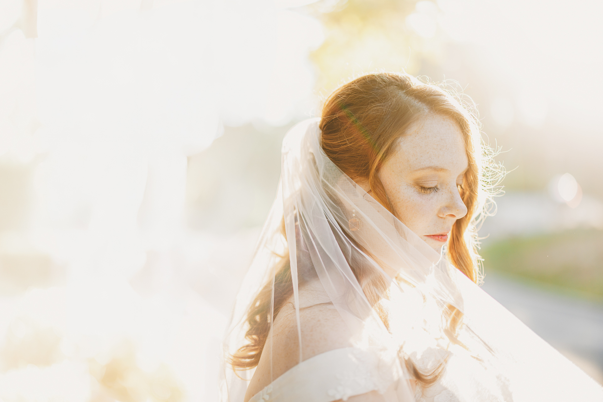 A bride with long hair, wearing a veil and an off-shoulder gown, looks down with a serene expression in bright, ethereal sunlight, almost as if she were an angel drafted straight from a dream.