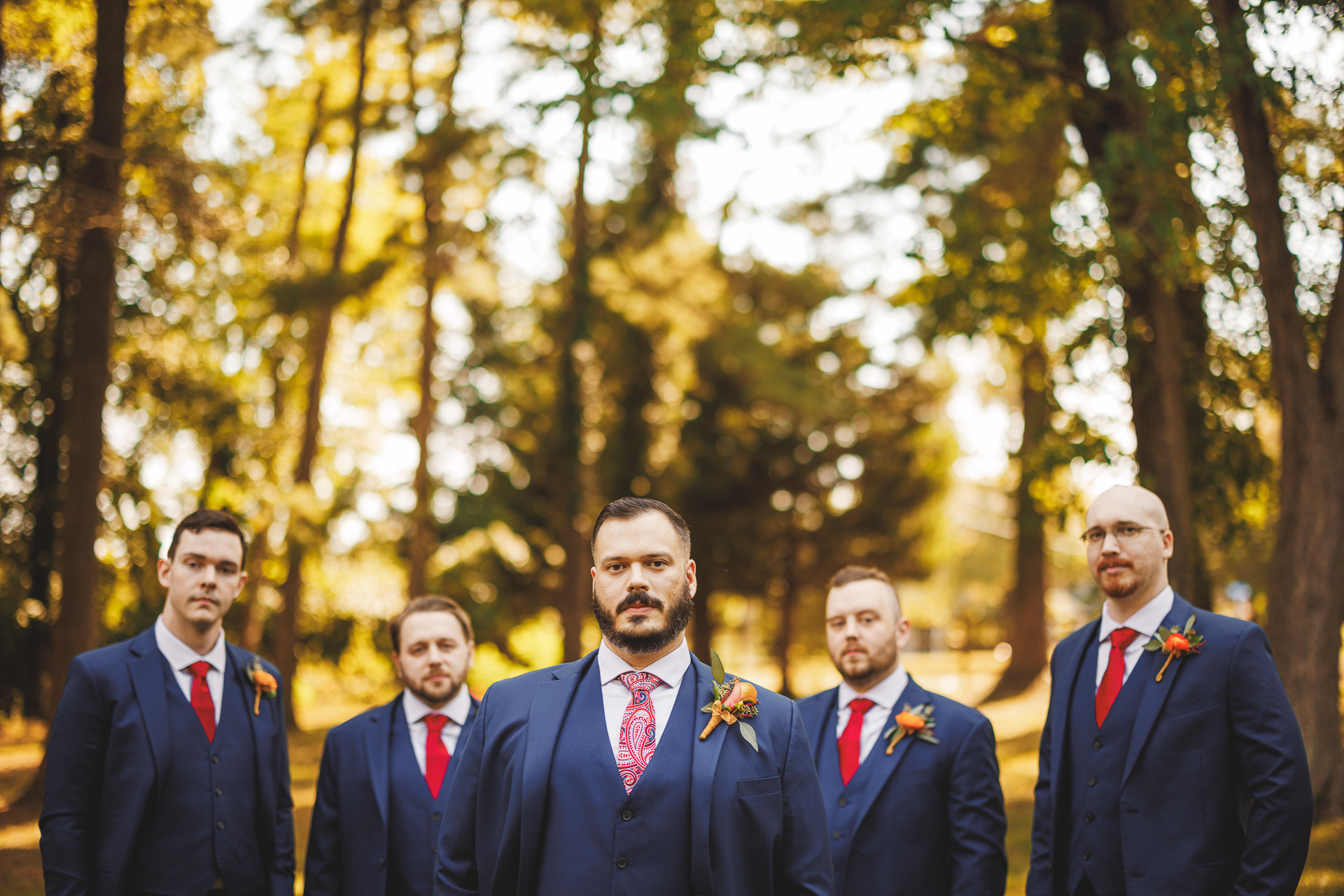 A group of five men dressed in formal suits with red ties and boutonnieres stand outdoors, with trees and sunlight in the background, looking like a carefully organized Auto Draft.