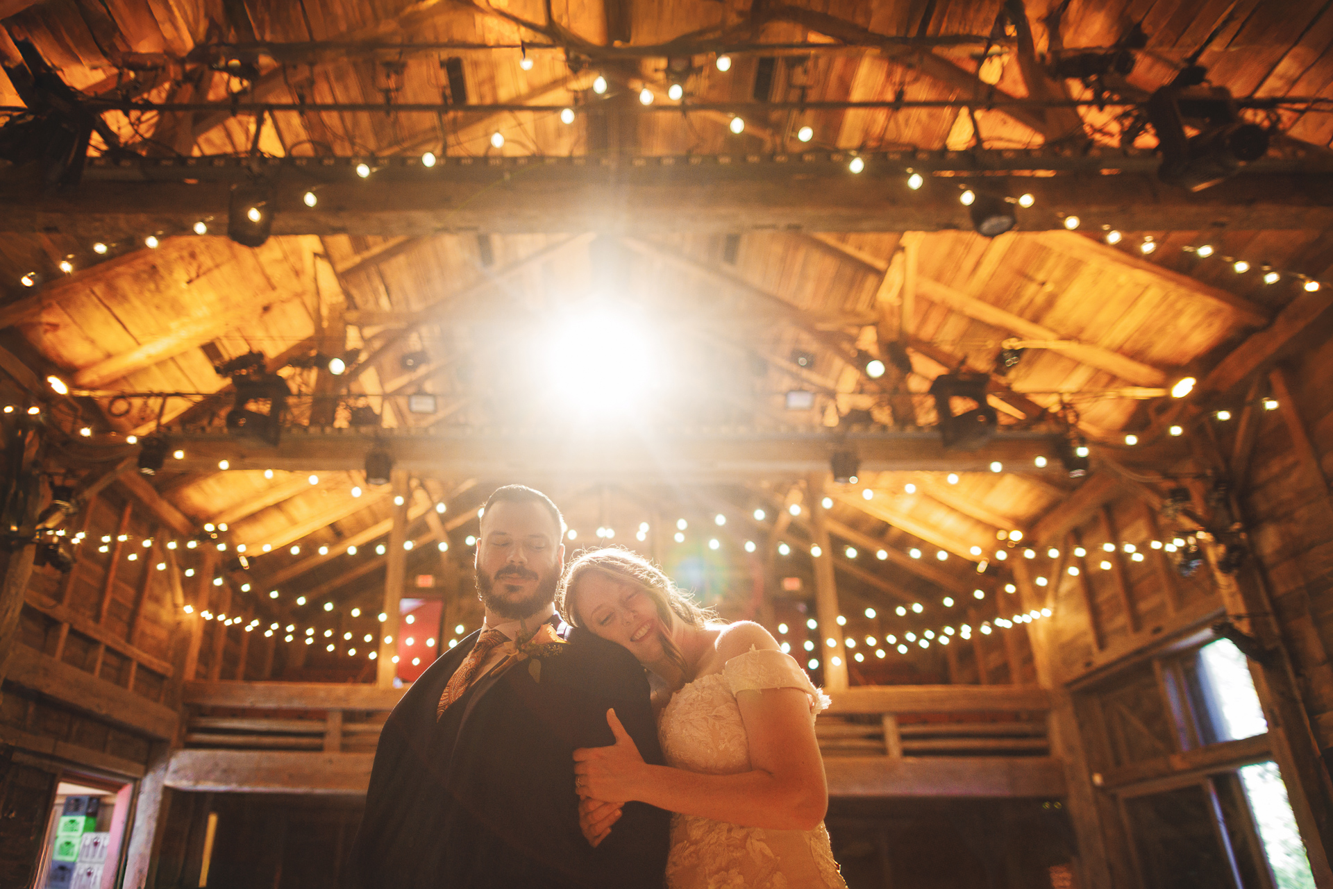 A couple poses under string lights inside a rustic wooden building. The woman rests her head on the man's shoulder as light rays from above illuminate the scene, capturing an auto draft of pure romance and tranquility.