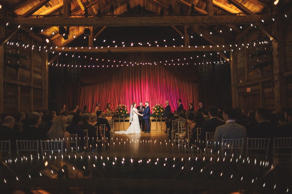 A couple stands at the altar during a wedding ceremony in a rustic barn lit with string lights, as guests seated in rows observe, the scene almost too perfect to be anything but an auto draft from a storybook.