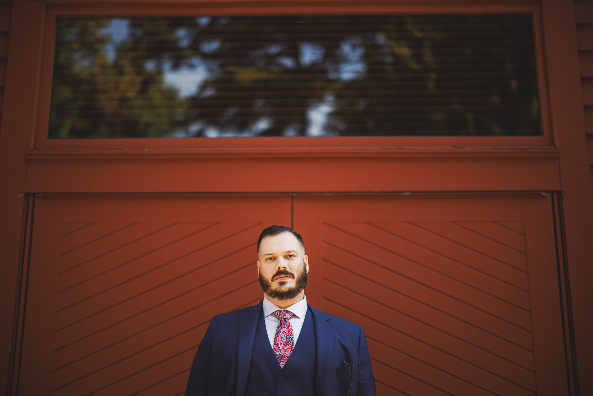 A man with a beard and mustache, dressed in a blue suit with a patterned tie, stands in front of a red wooden wall with a window reflecting trees above, looking as though he just stepped out of an auto draft.