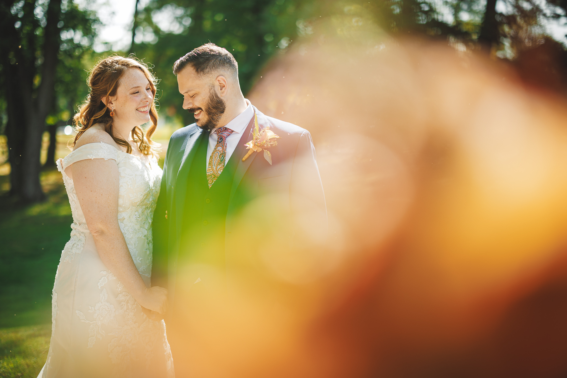 A bride and groom stand closely together outdoors, smiling at each other. The background is blurred with sunlight and greenery, creating a warm, glowing effect around them, like an auto draft capturing nature's perfect moment.