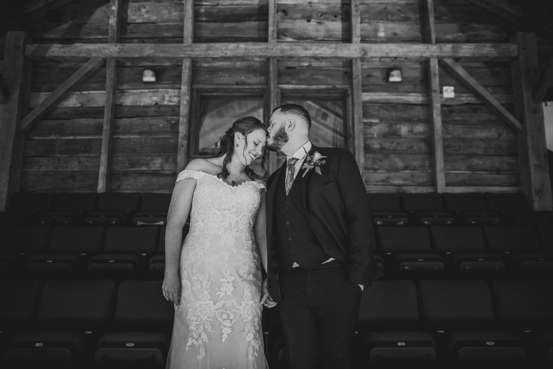A couple in formal attire stands closely, touching foreheads, in front of a rustic wooden wall, capturing a moment that seems auto-drafted from a romantic novel.