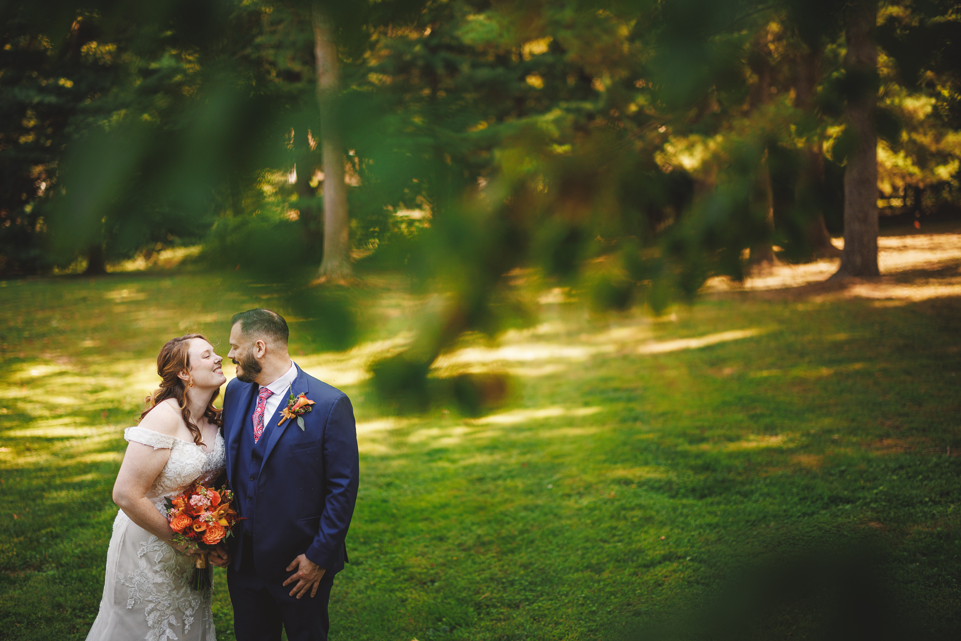 A bride in a white dress and groom in a blue suit share a kiss in a grassy, wooded park with sunlight filtering through the trees, creating an enchanting moment that seems almost like it was captured for an auto draft.