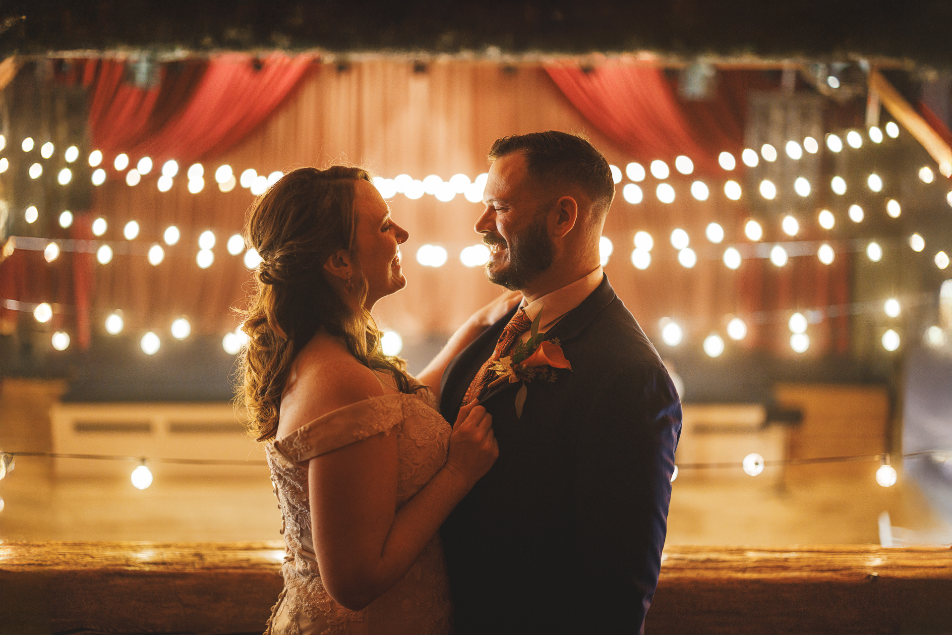 A couple dressed in wedding attire stands close, smiling at each other, with string lights and draped curtains in the background creating a romantic atmosphere reminiscent of a picture-perfect auto draft.