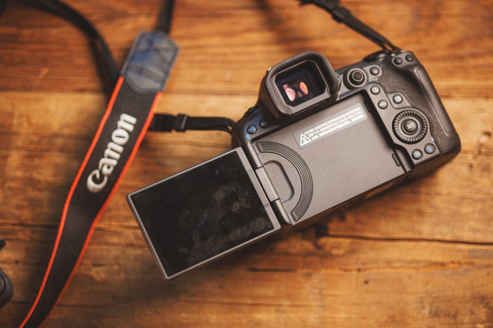 A Canon R5 Mark ii DSLR camera with an open LCD screen rests on a wooden surface. The camera strap is visible on the left, showcasing its practicality for real-world use.