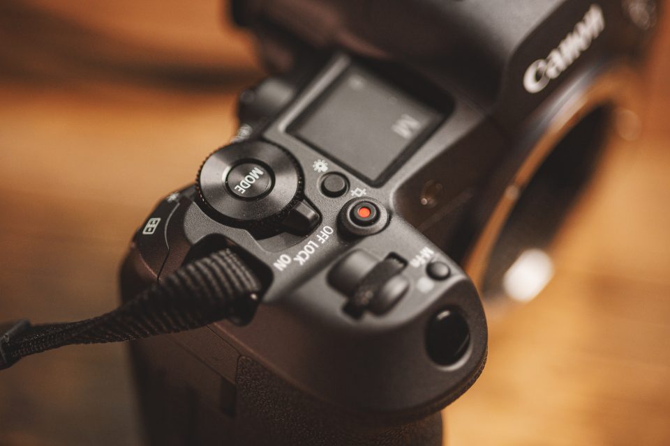 Close-up of a Canon camera, focusing on the mode dial, record button, and various control buttons. The camera has a black strap attached.
