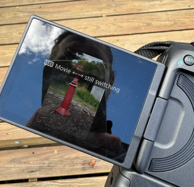 Close-up of a DSLR camera screen displaying a reflection of a red hydrant with the text "Movie still switching." The camera is resting on a wooden surface, highlighting real-world use. This could easily be part of a Canon R5 Mark ii Review, demonstrating its impressive capabilities.