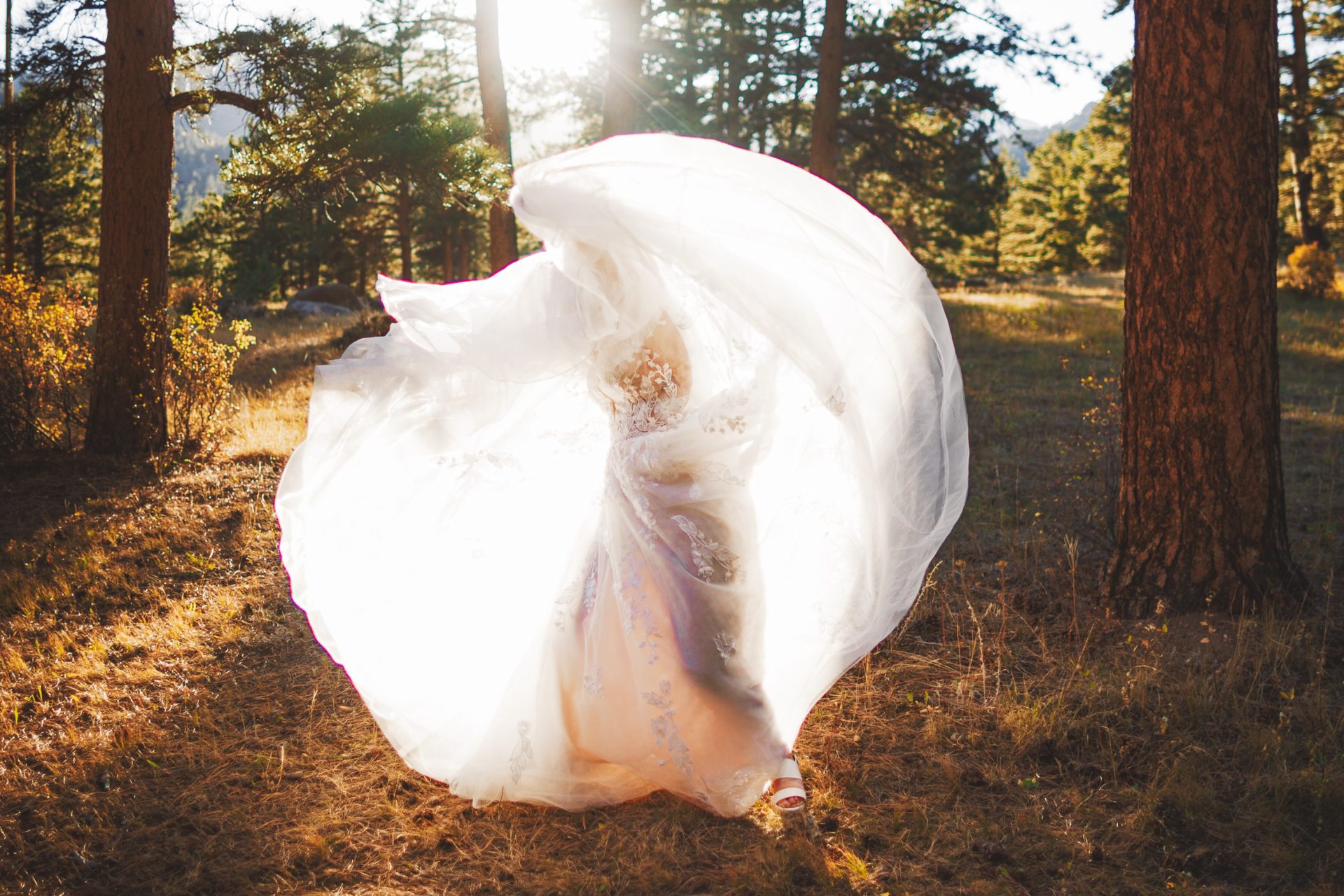 A person in a flowing white dress stands in a sunlit forest clearing at Della Terra Mountain Chateau, with the fabric billowing around them.
