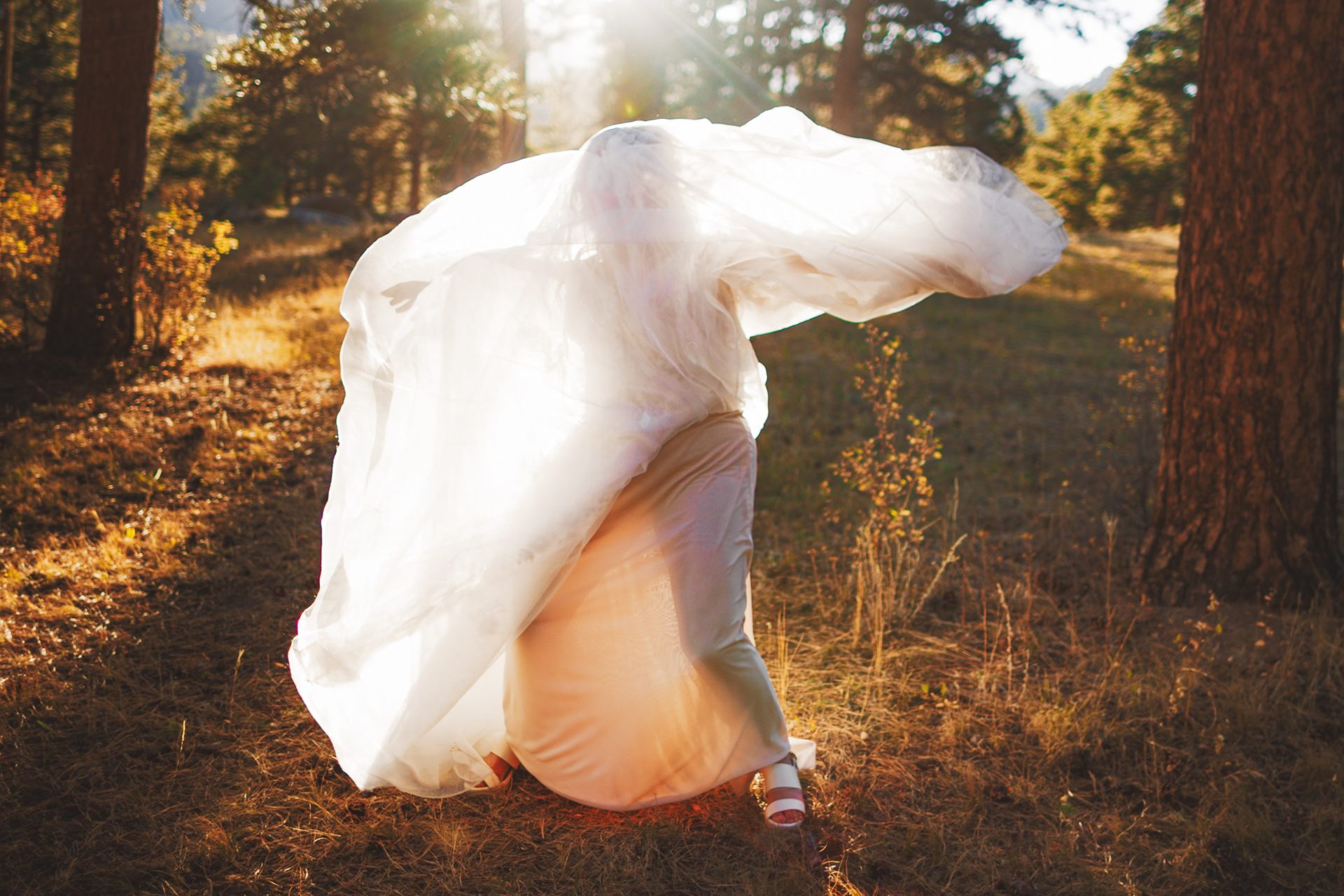 A person crouches in a forest clearing, covered by a billowing white fabric, with sunlight streaming through the trees.
