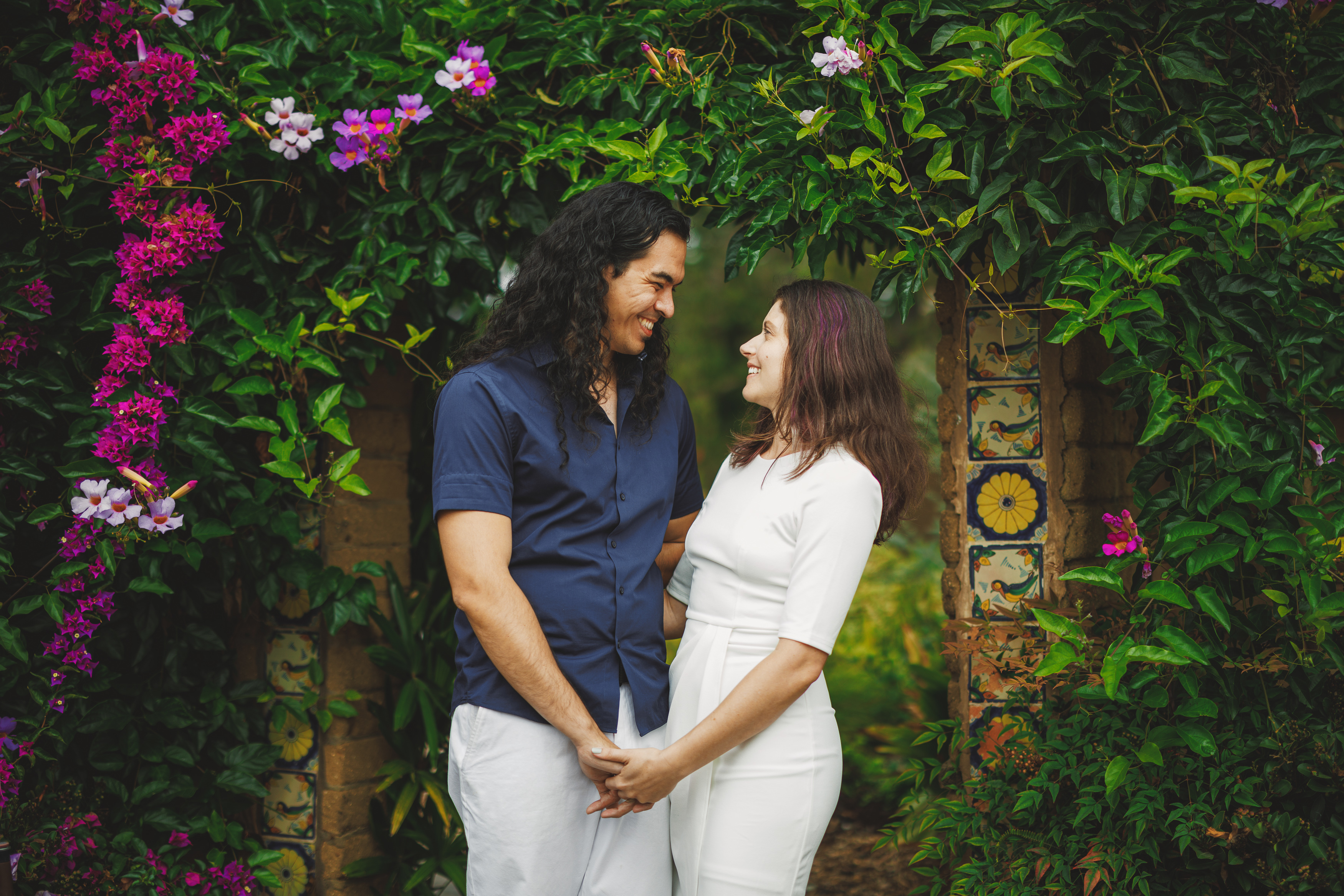 A couple stands holding hands, smiling at each other under an archway adorned with colorful flowers and lush greenery, perfectly capturing a moment in the enchanting San Francisco Botanical Garden.