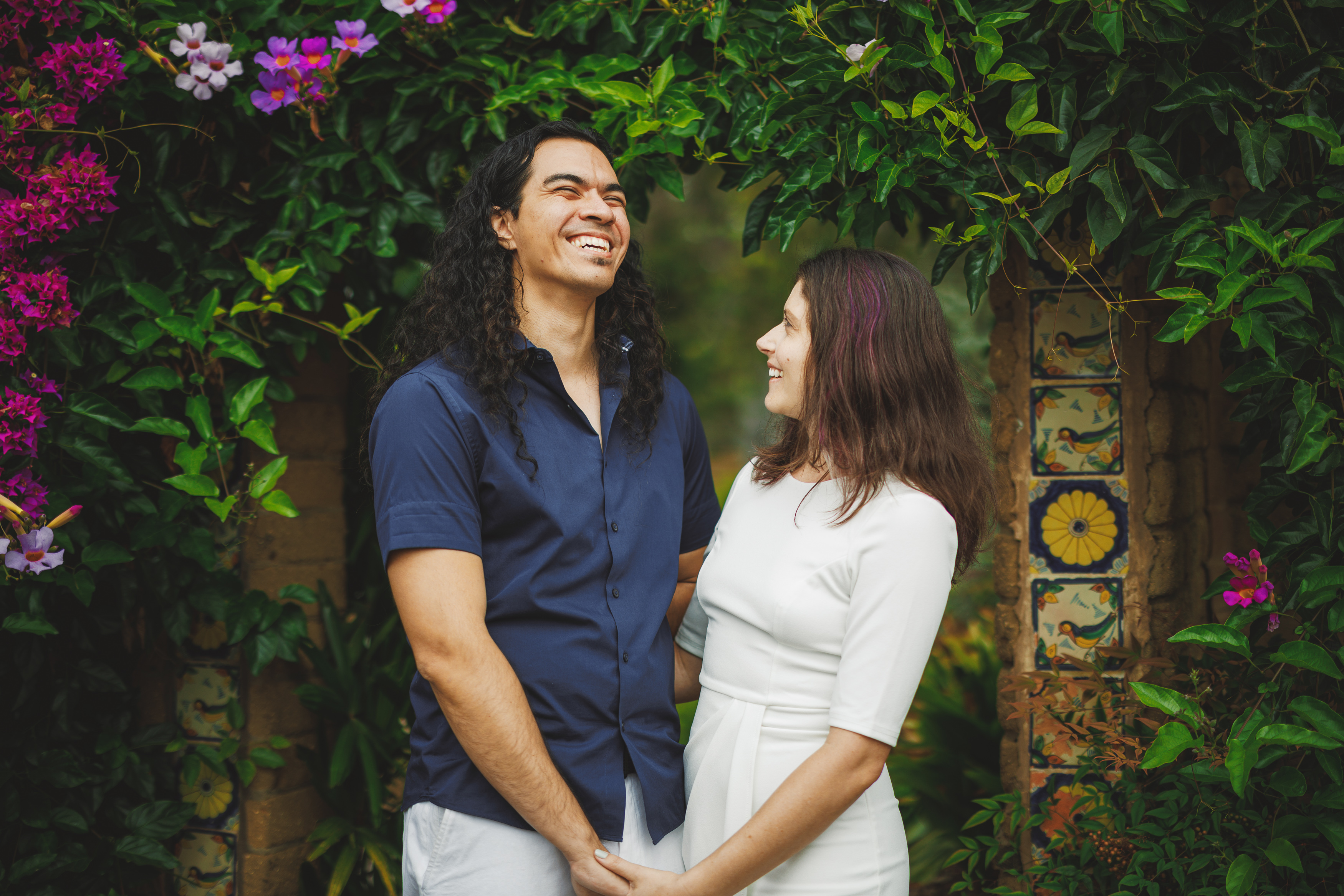 A couple stands smiling under an archway adorned with vibrant pink flowers and greenery, holding hands.