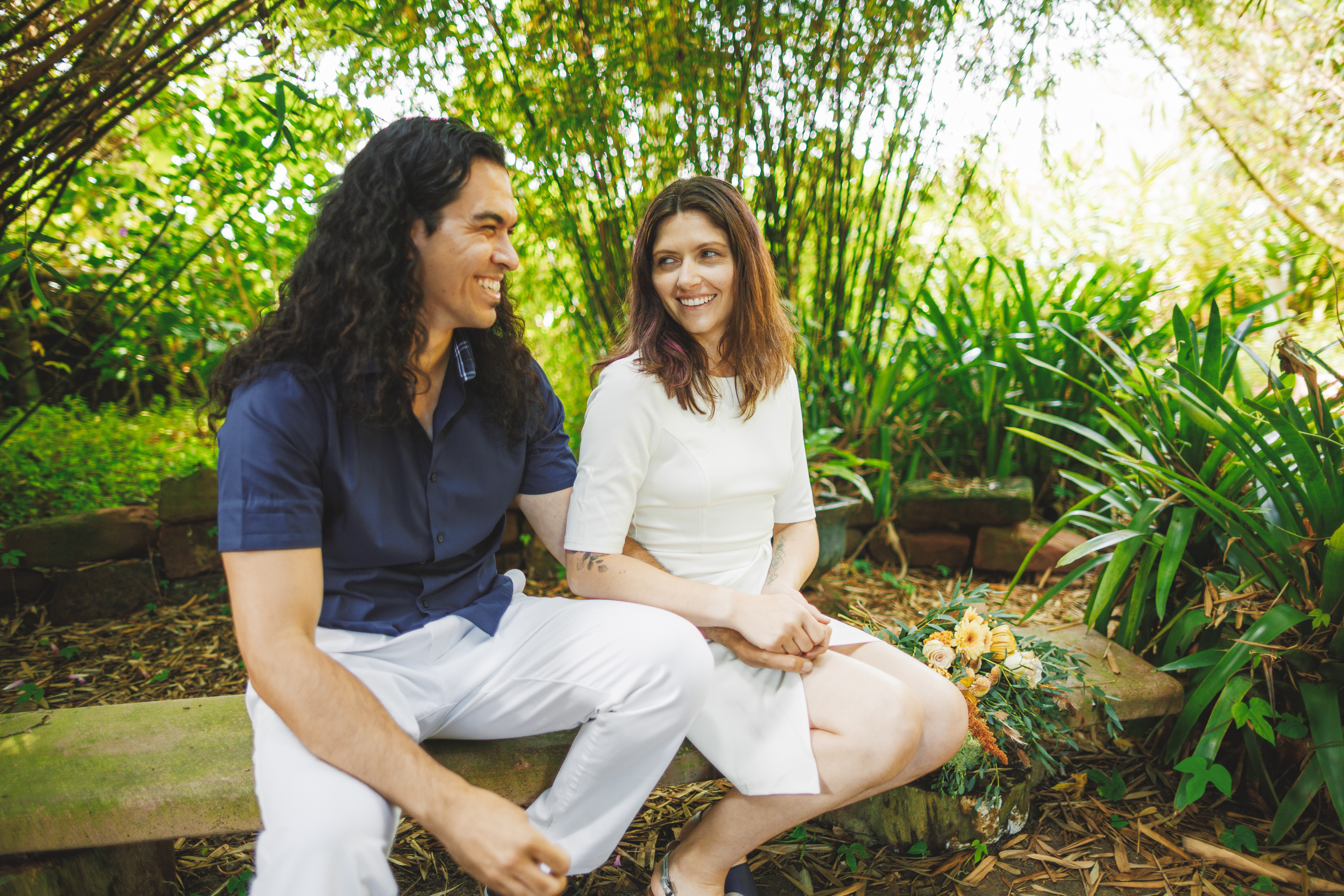 A couple sits on a bench in a garden, smiling at each other. A bouquet of flowers rests beside them.