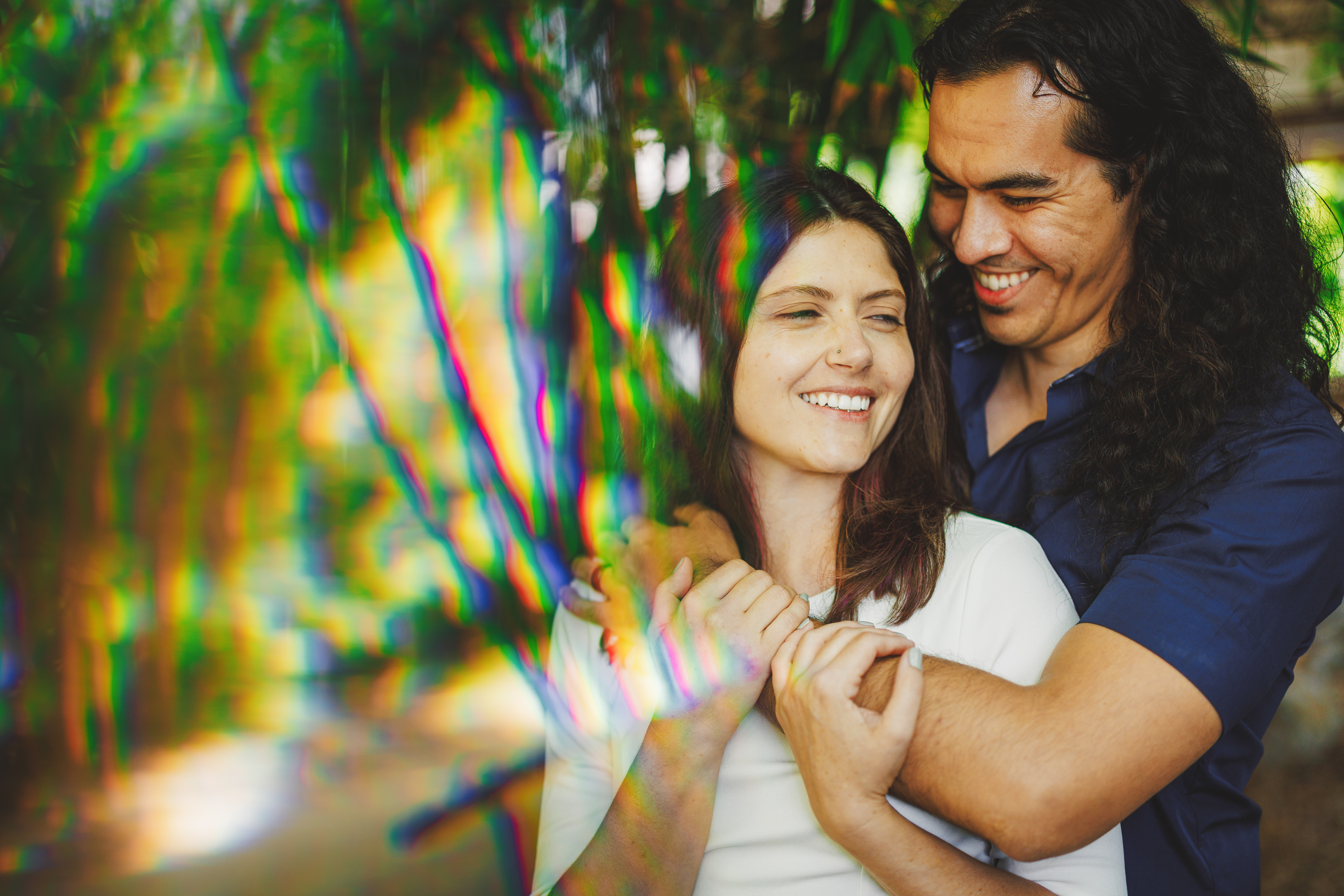 A smiling man and woman embrace outdoors, surrounded by a colorful, rainbow-like effect and greenery.
