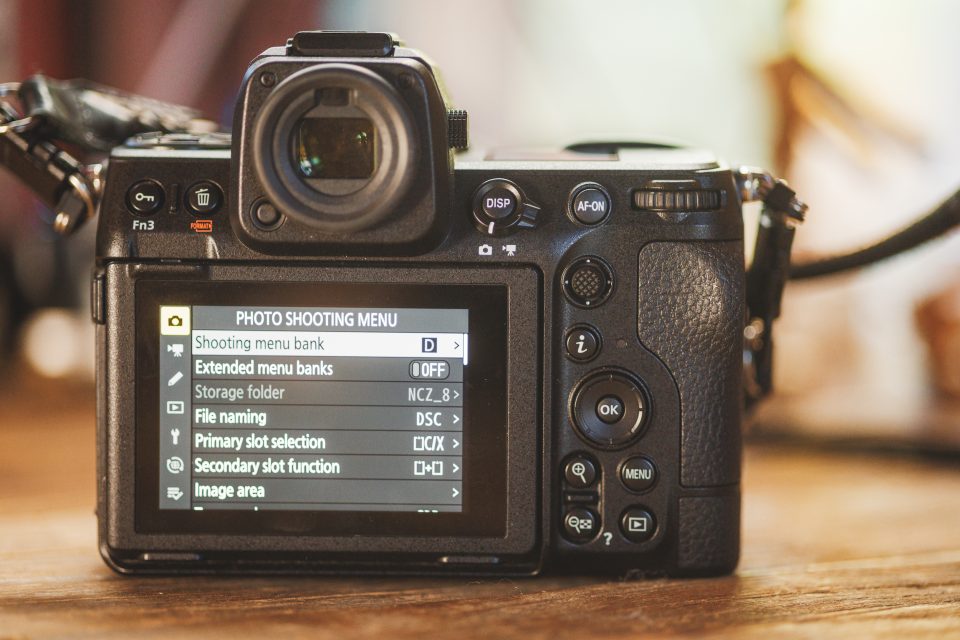 A digital camera rests on a wooden surface, with the photo shooting menu and Auto Draft options prominently displayed on its screen.