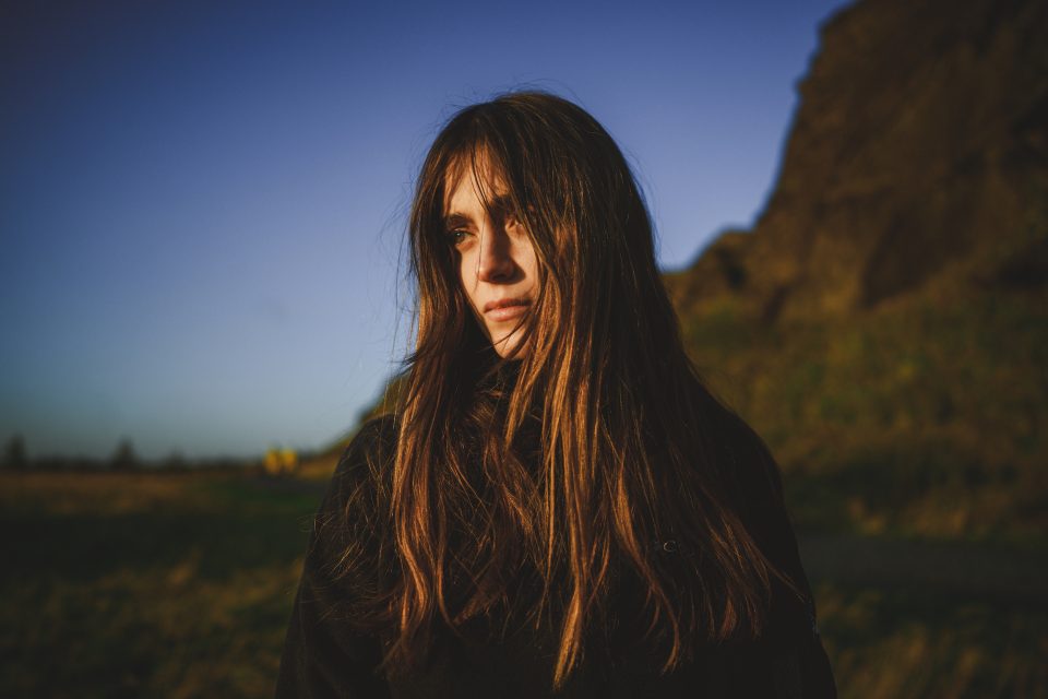 A person with long brown hair stands outdoors, facing sideways, their silhouette almost like a serene auto draft under the sun's glow. The background features a clear sky and a grassy area, capturing a moment of natural beauty.