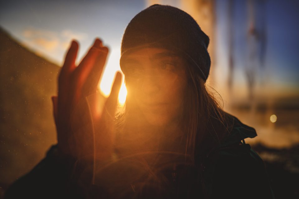 A person wearing a beanie is bathed in the sun's glow, their hand reaching towards the light. The auto-draft of nature unfolds behind them with a majestic waterfall set against a stunning sunset.